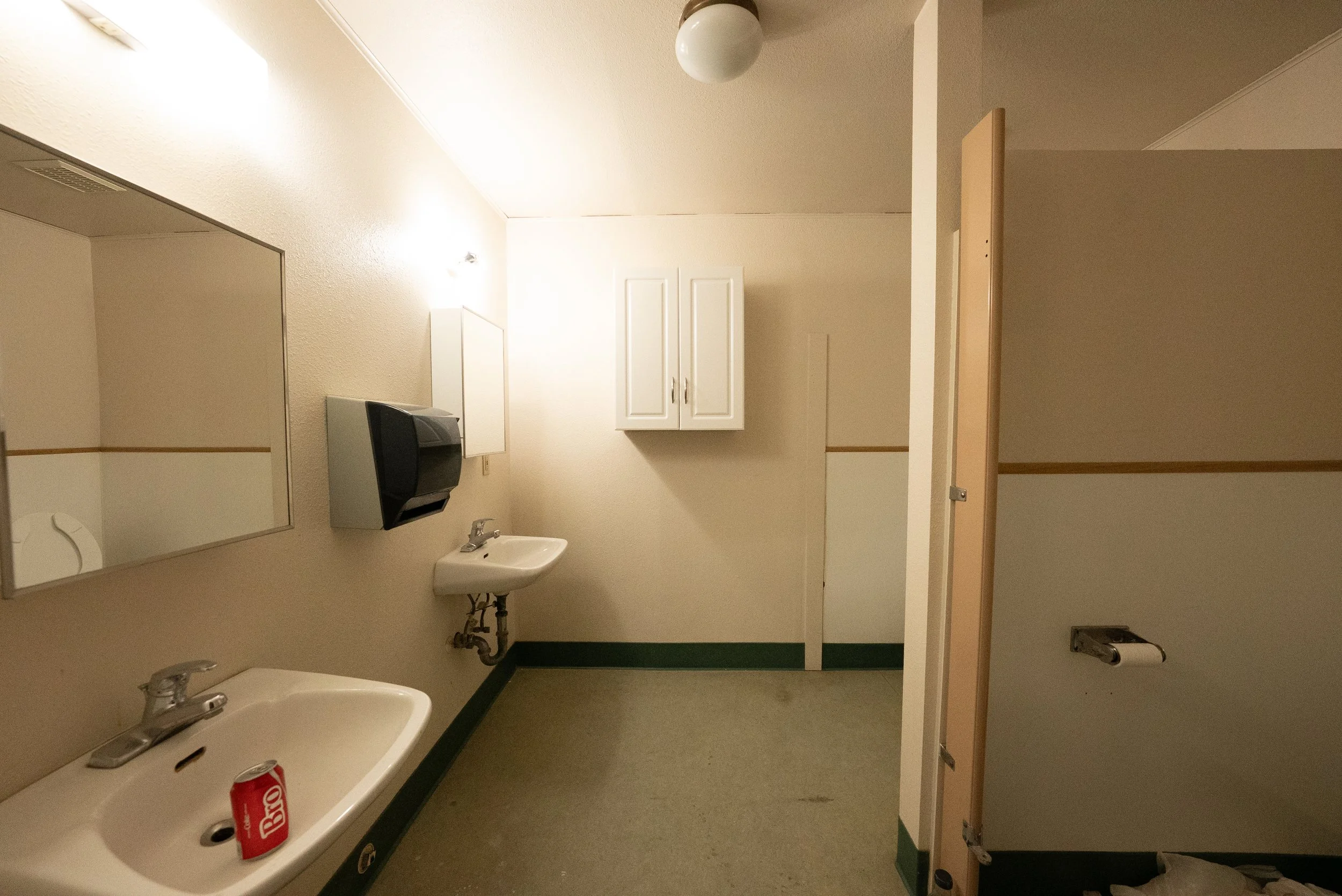 A hospital or clinic bathroom with two sinks, a mirror, a small medicine cabinet, a paper towel dispenser, and a door partially open revealing a stall. The bathroom has beige walls, a green and beige floor, and bright lighting.