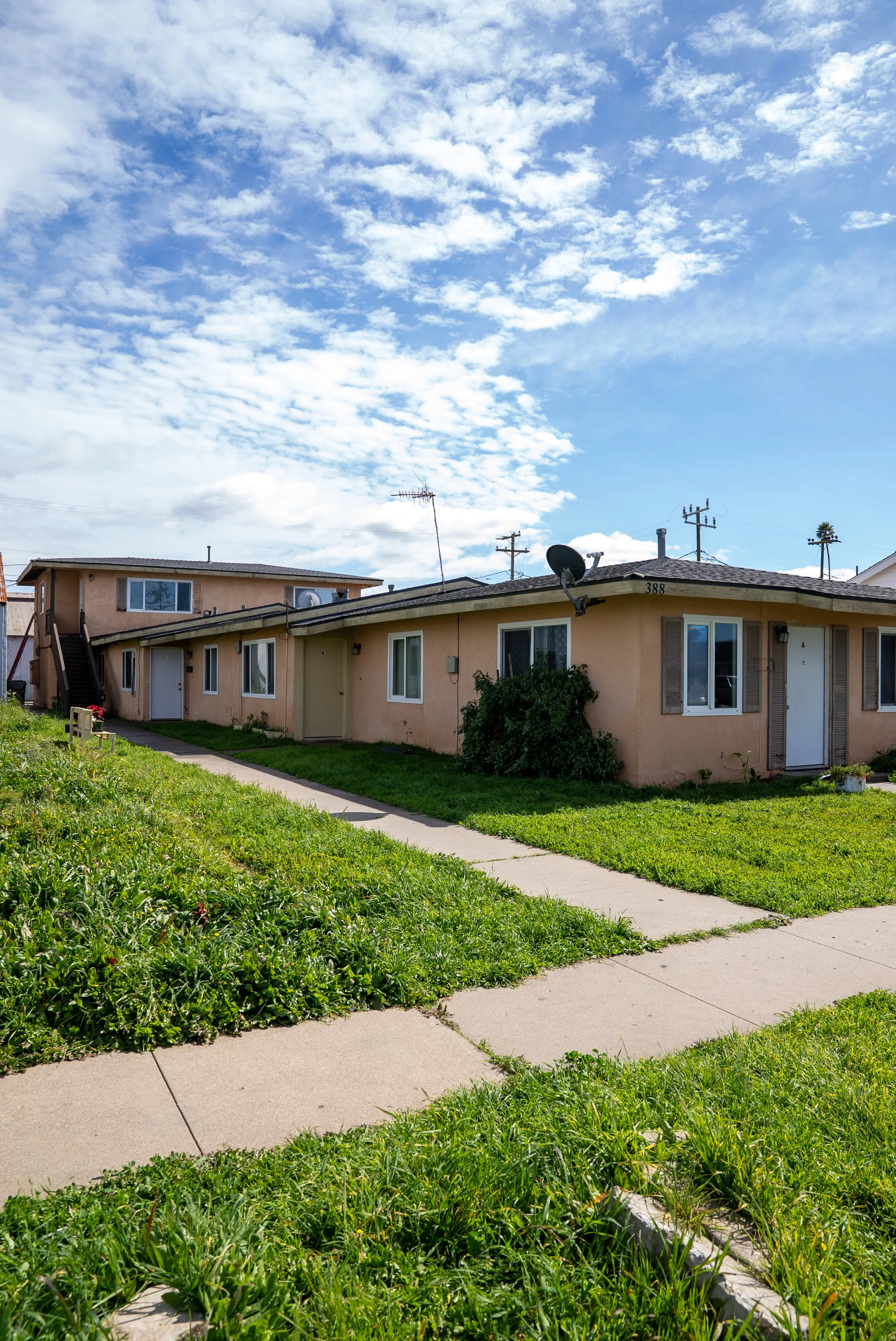 A residential area with a beige apartment building, a grassy lawn, a concrete sidewalk, and a bright blue sky with scattered clouds.