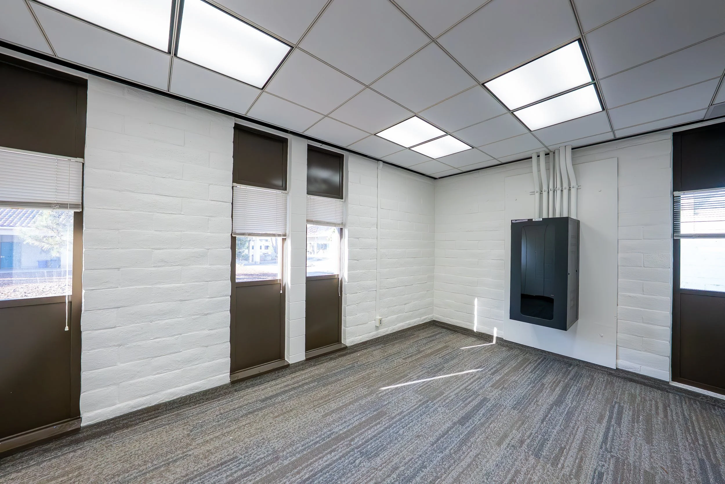 Empty office room with white brick walls, windows with blinds, gray carpet, and ceiling lights.