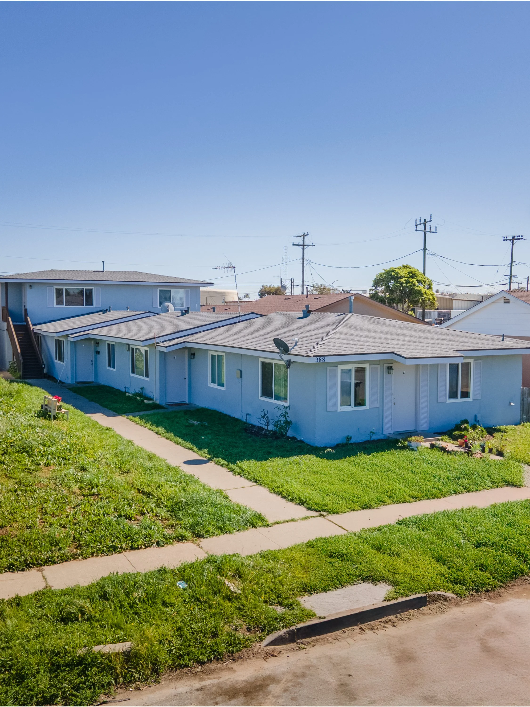 A view of a neighborhood with several houses under a clear blue sky, with green lawns, sidewalks, and power lines in the background.
