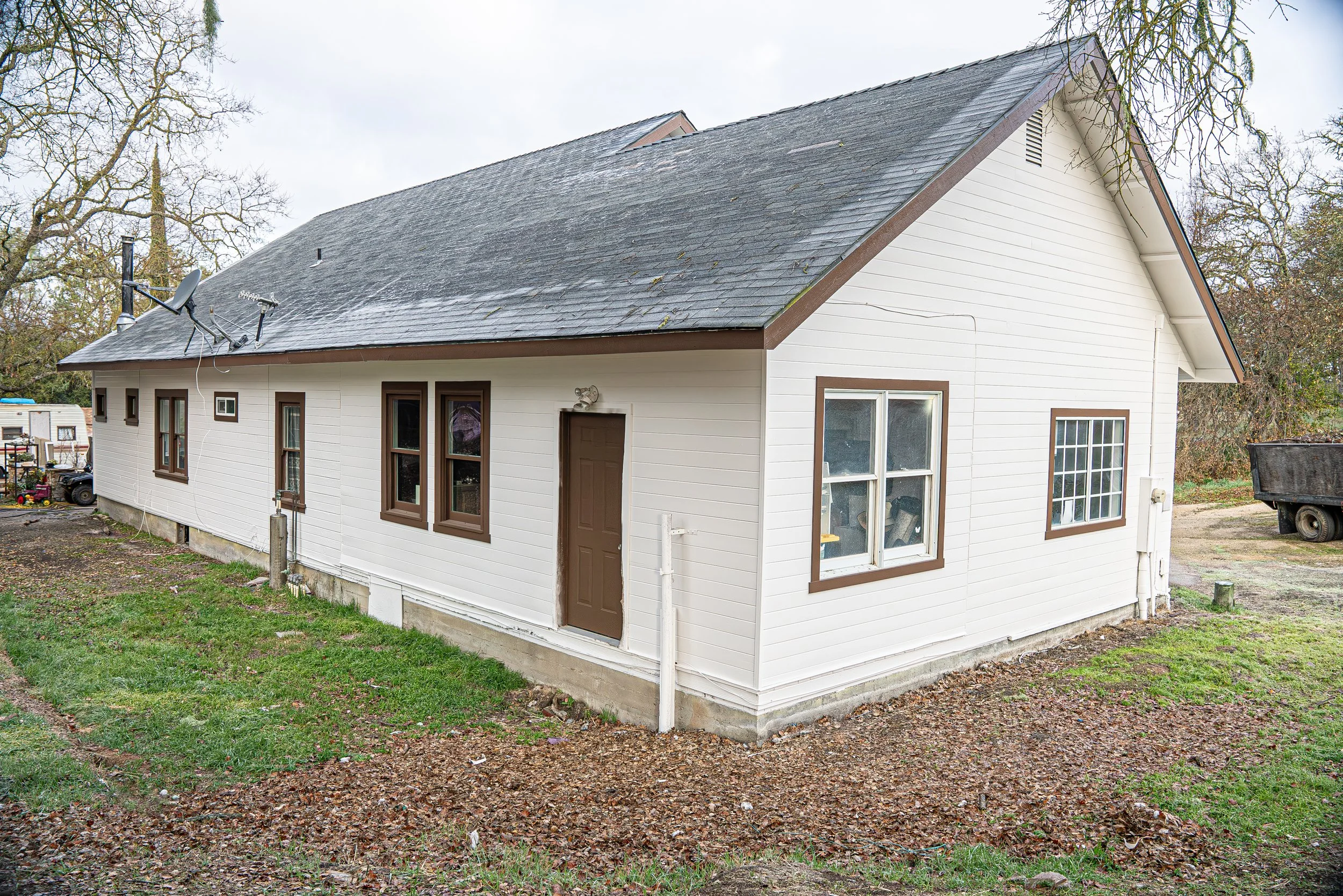 A white house with brown window frames and brown door, sitting on a raised concrete foundation, with a gray shingle roof and a satellite dish attached. The yard has patches of grass and dirt, and trees with mostly bare branches surround the house.