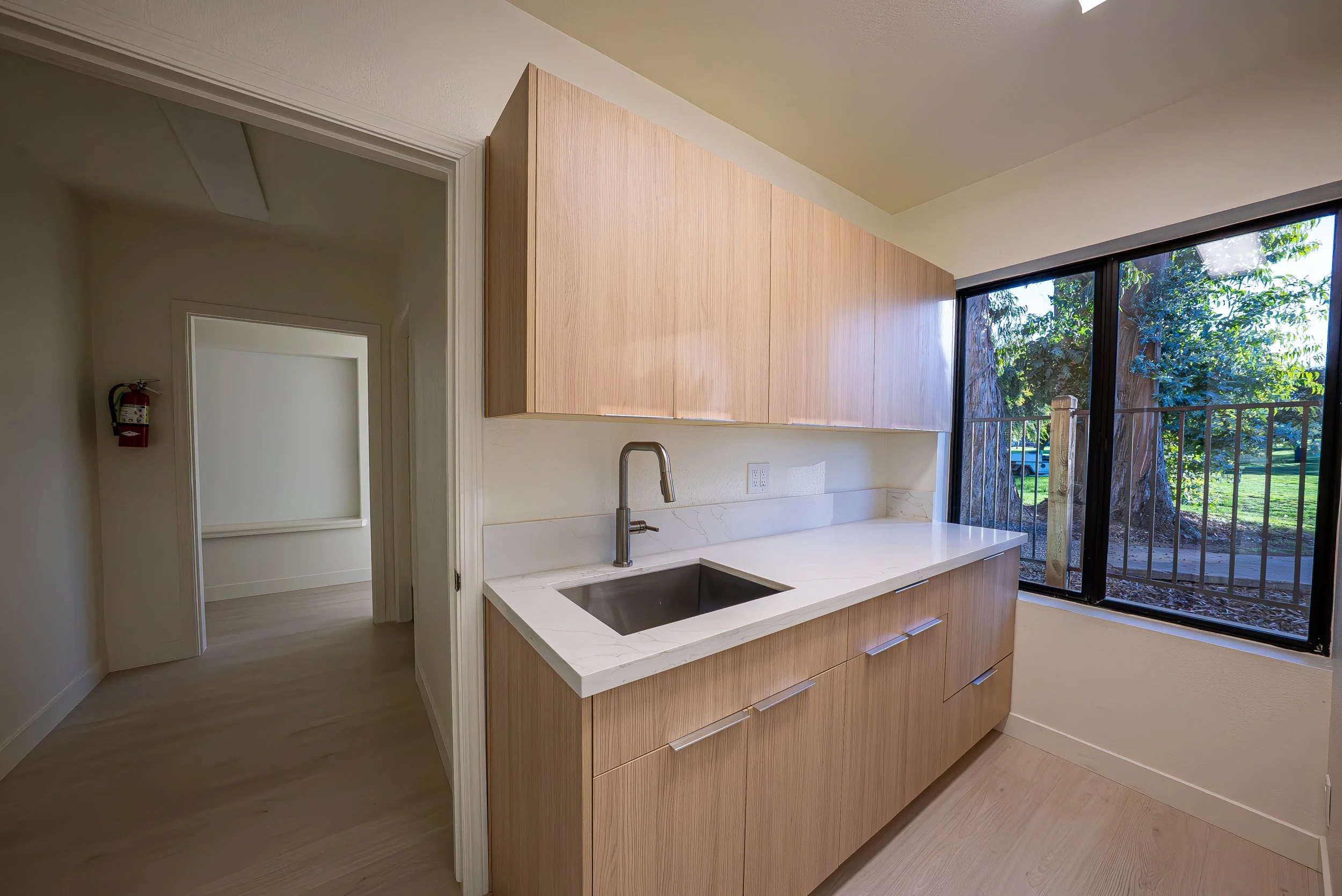 Kitchen with light wood cabinets, a white marble countertop, a stainless steel sink, and large windows showing trees outside.