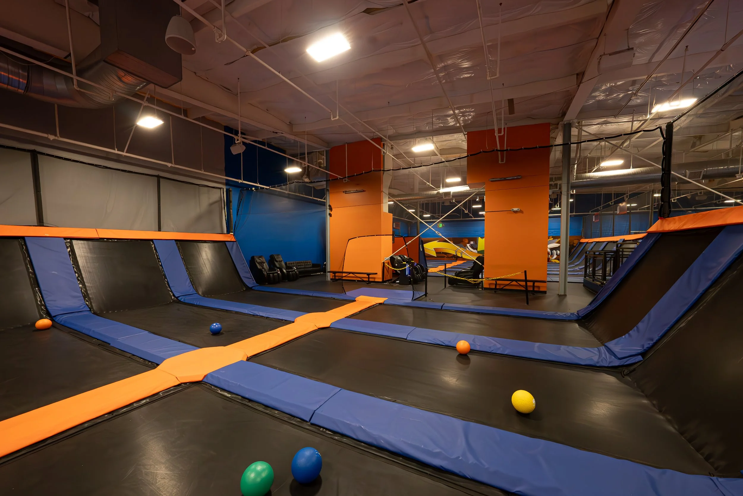 Indoor trampoline park with multiple trampolines, colorful balls, orange padded edges, and a colorful wall in the background.