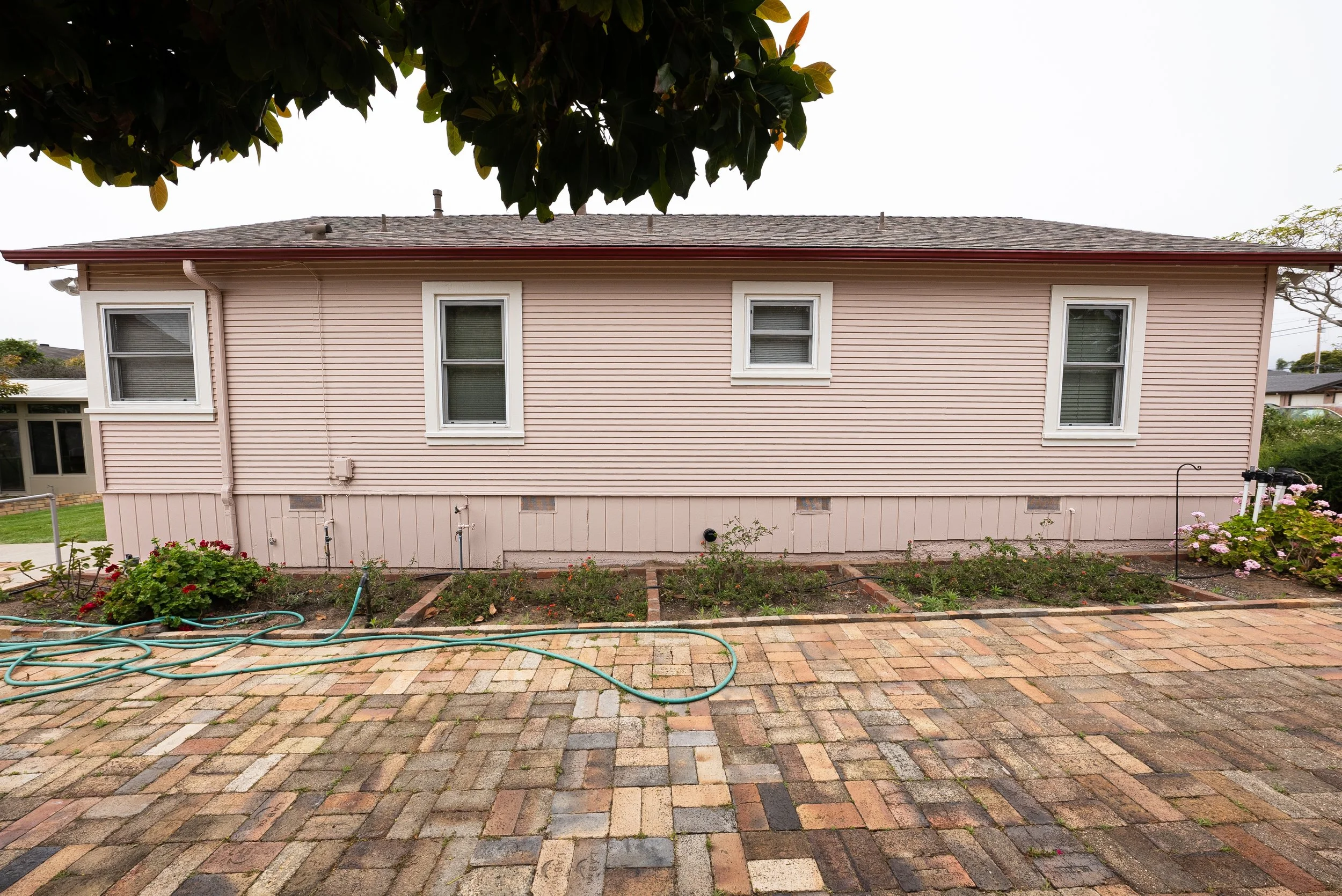 Backyard view of a pink house with three windows, a garden with plants and flowers, a brick patio, and a cloudy sky overhead.