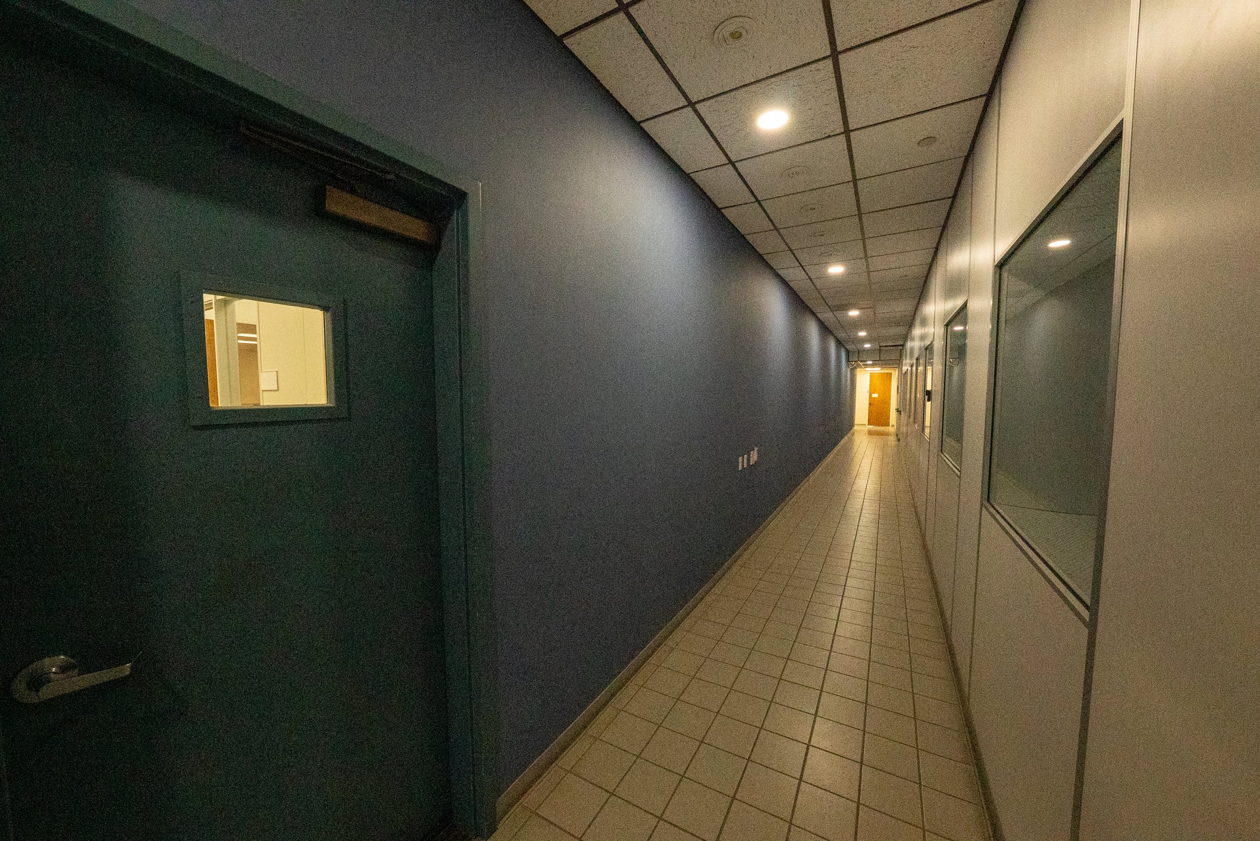 A hallway in a building with a dark wall on the left, multiple ceiling lights, tiled floor, and glass doors on the right side.