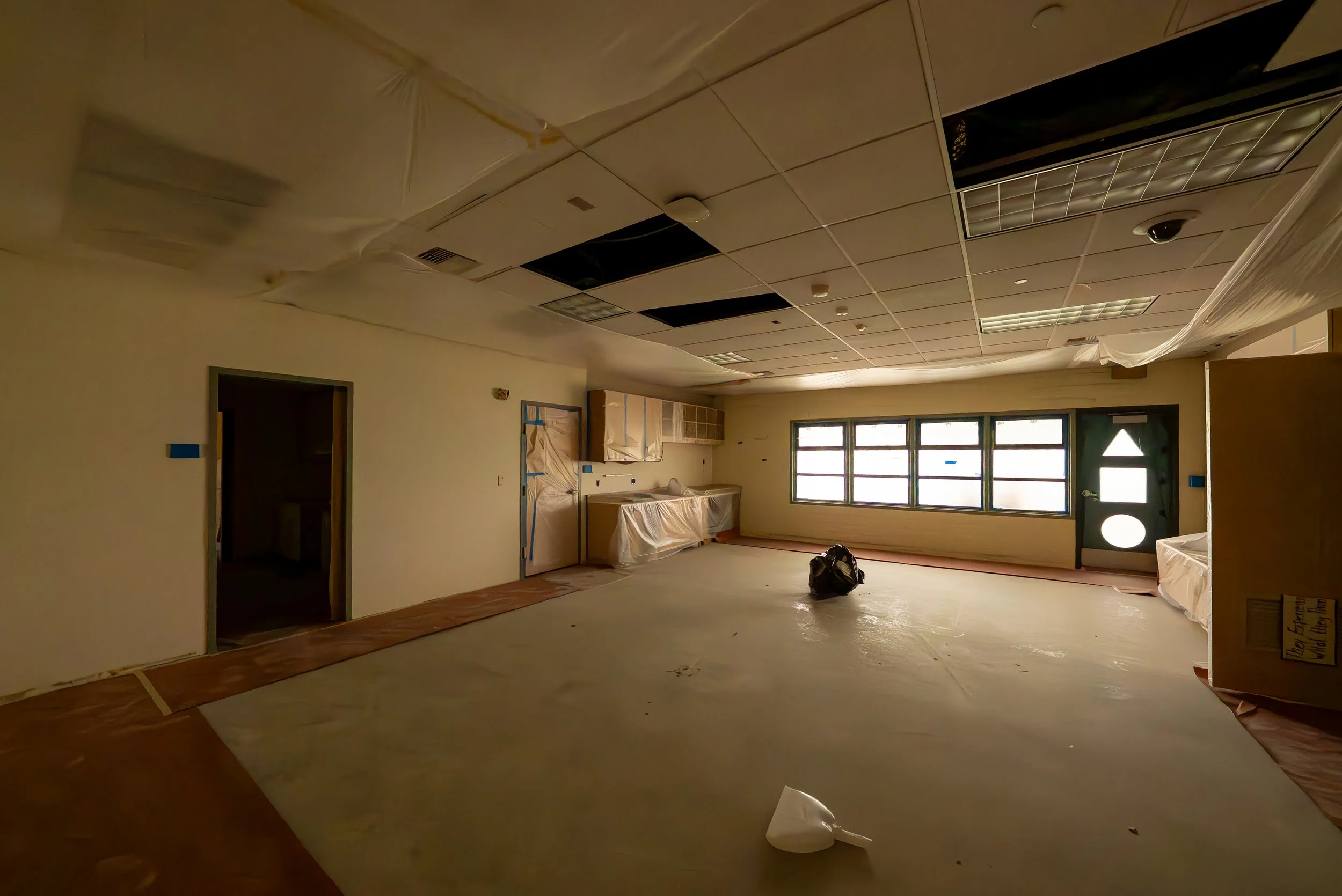 Empty room under renovation with ceiling tiles removed, plastic coverings, and construction materials, illuminated by natural light through large windows.