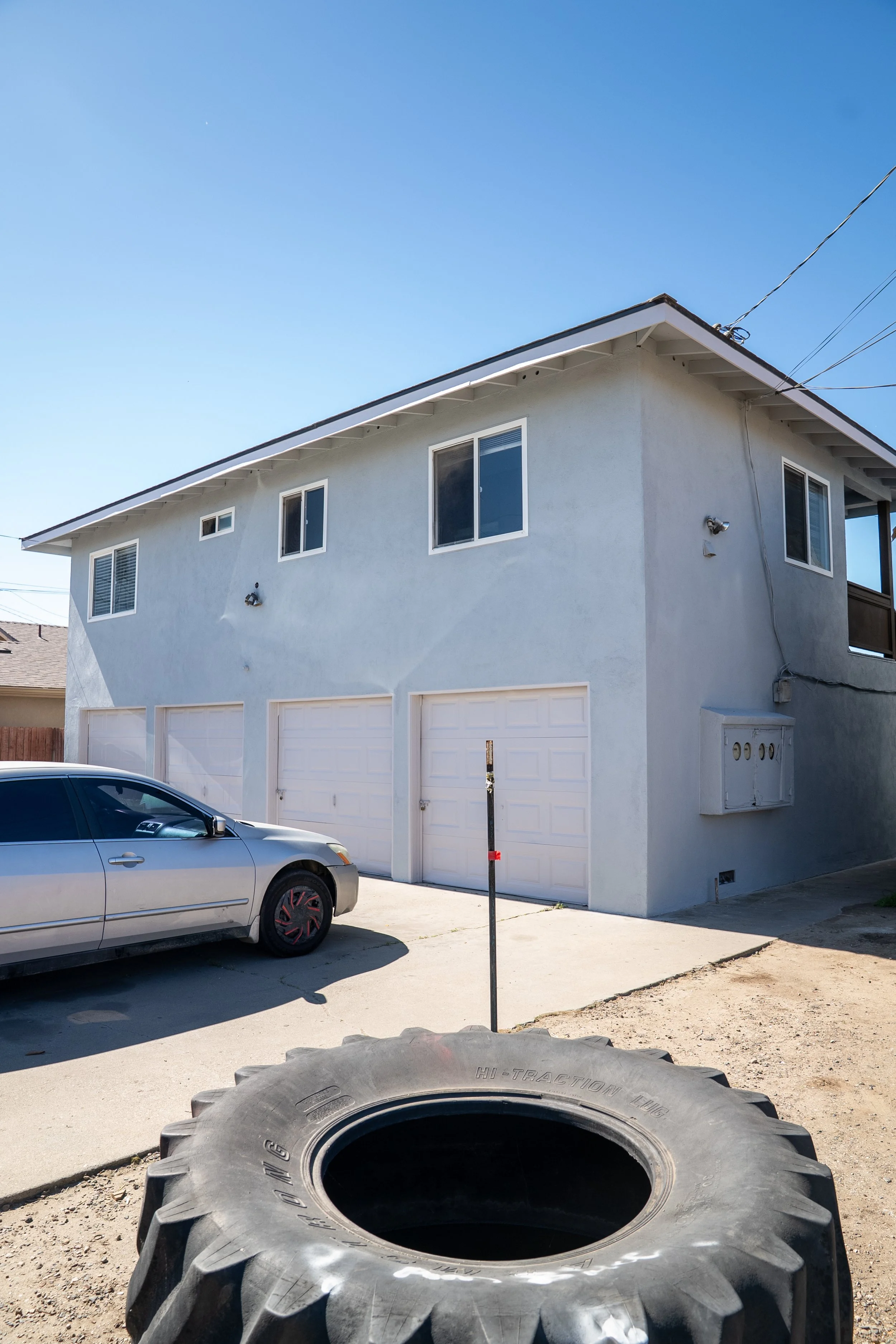 A two-story light blue house with multiple windows and a garage, in front of which is a silver car parked. Large tires in the foreground and a blue sky above.