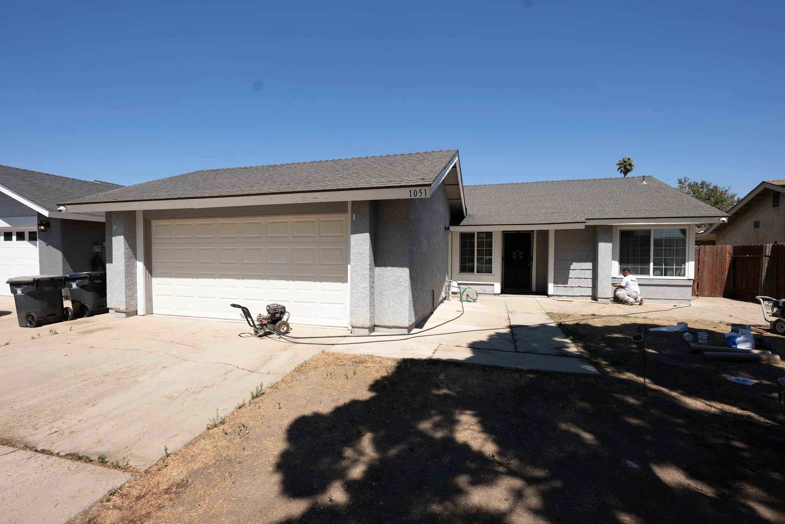 A single-story house with a gray roof, white garage door, and a concrete driveway. A man is working on the house's exterior wall. There are trash cans and various tools in the driveway. Clear blue sky and a palm tree are visible in the background.