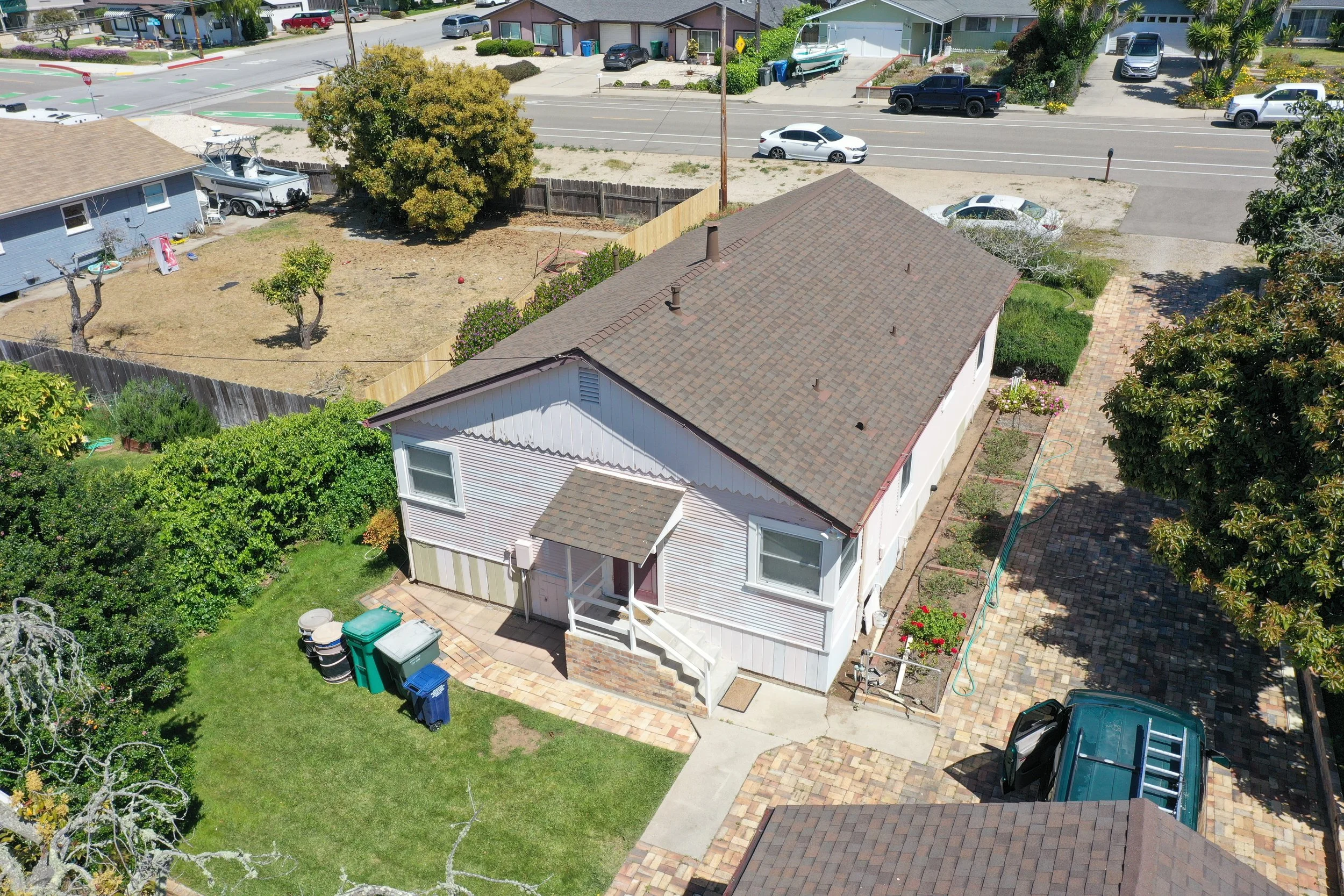 An aerial view of a suburban backyard with a house, a grassy lawn, trees, a paved patio, and various garbage bins. The backyard is enclosed by a wooden fence, and neighboring houses and streets are visible in the background.