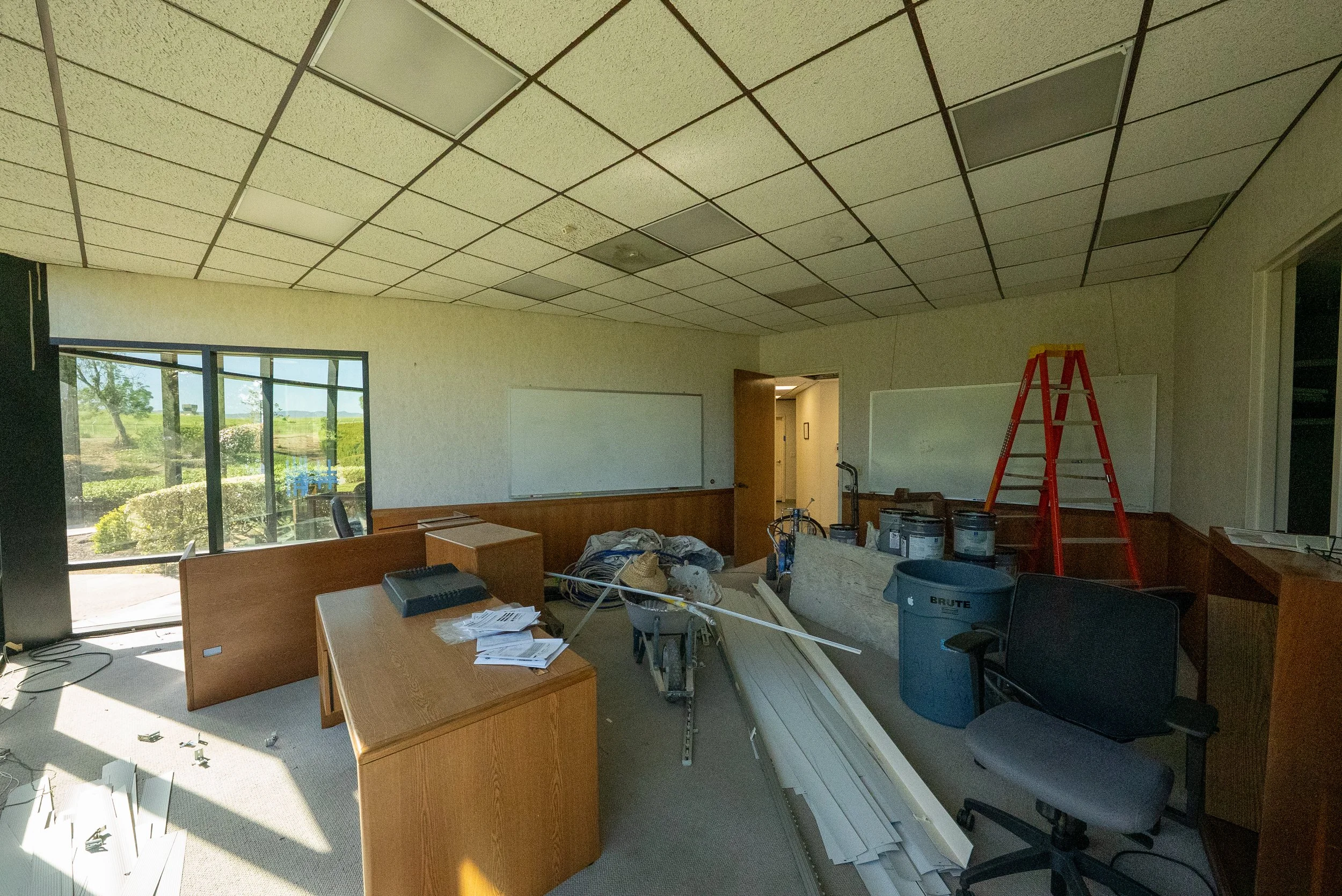 Office room under renovation, with cluttered desks, construction materials, and a red ladder, window view of trees outside.