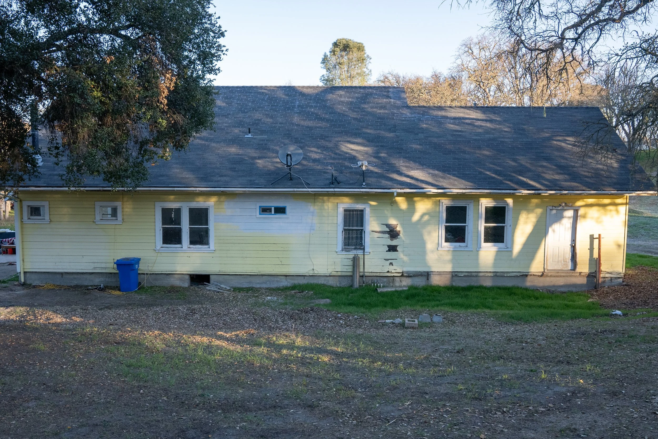 A yellow house with a grey shingled roof, four windows on the front, and a white door. The roof has two antennas mounted. There is a blue trash bin outside the house, and the yard has grass and dirt. Large trees with mostly bare branches surround the