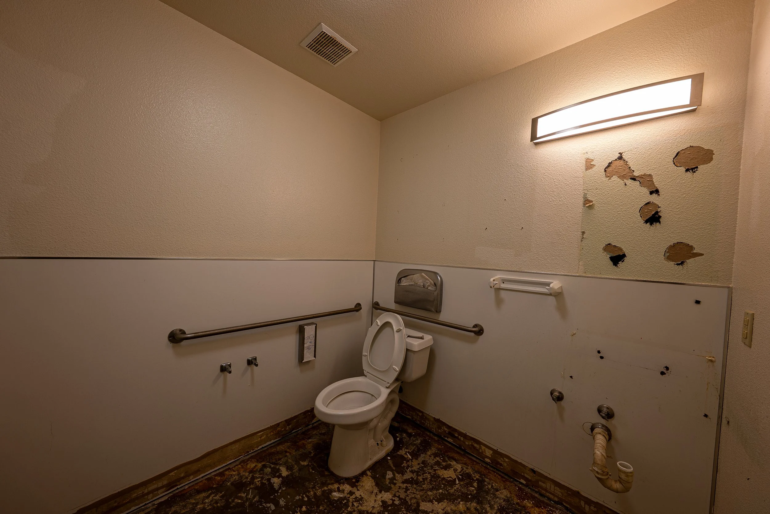 Restroom with a toilet, damaged wall with peeling paint, railings, and flooring.