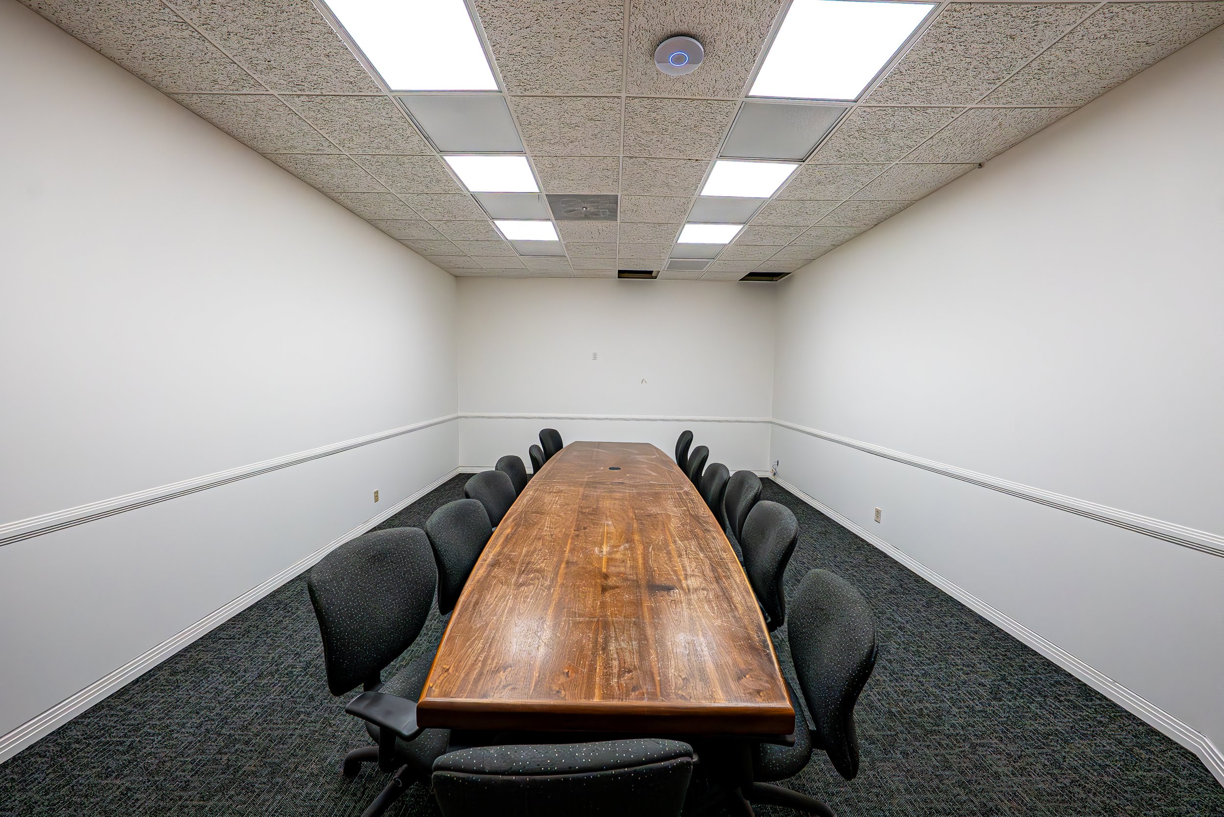 Empty conference room with a wooden table and black chairs surrounded by white walls, ceiling with fluorescent lights and ventilation panels.