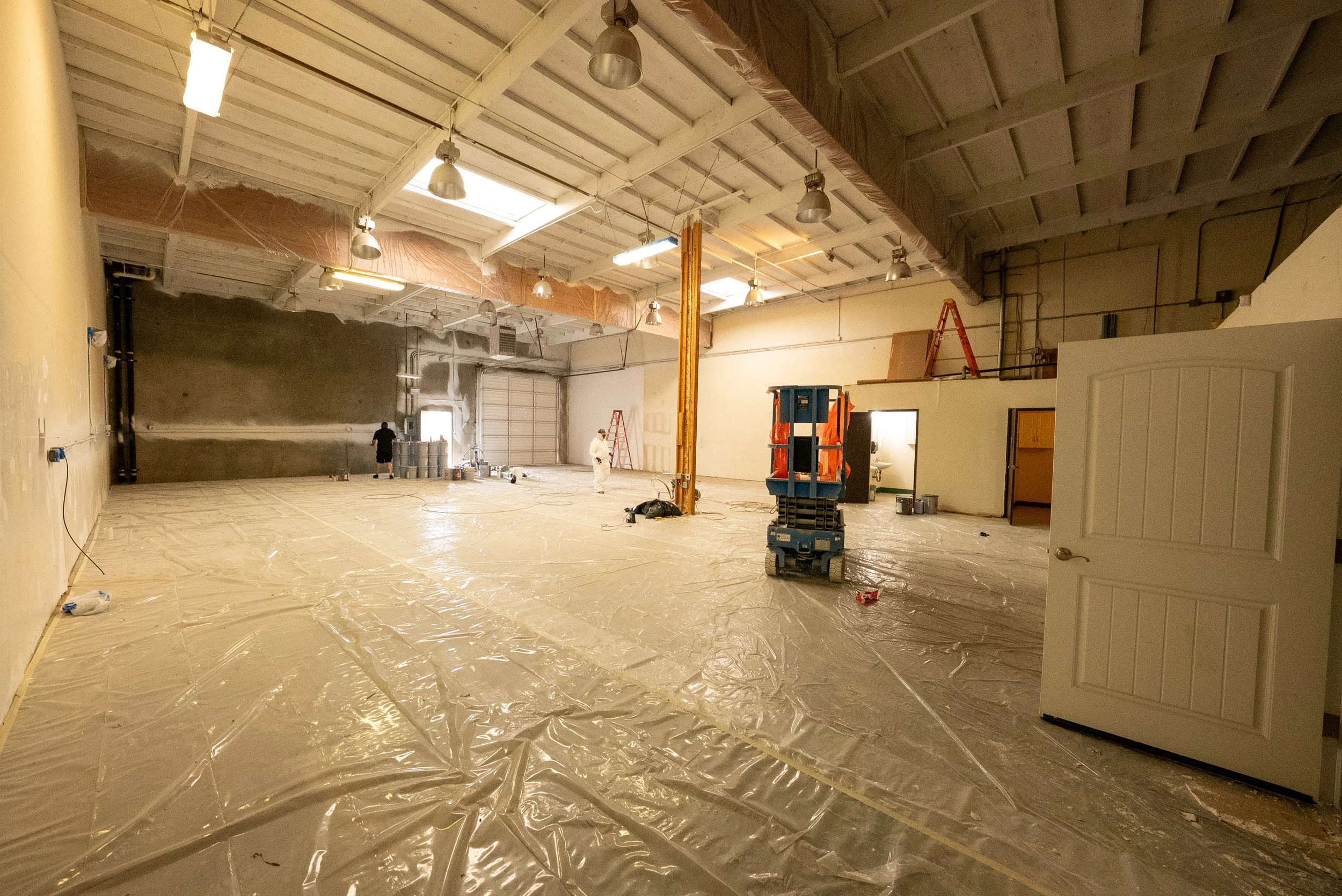 The image shows an indoor construction or renovation site with workers, construction equipment, and materials. The space has a high ceiling with multiple lights hanging from it. The floor is covered with a protective plastic sheet for construction wo