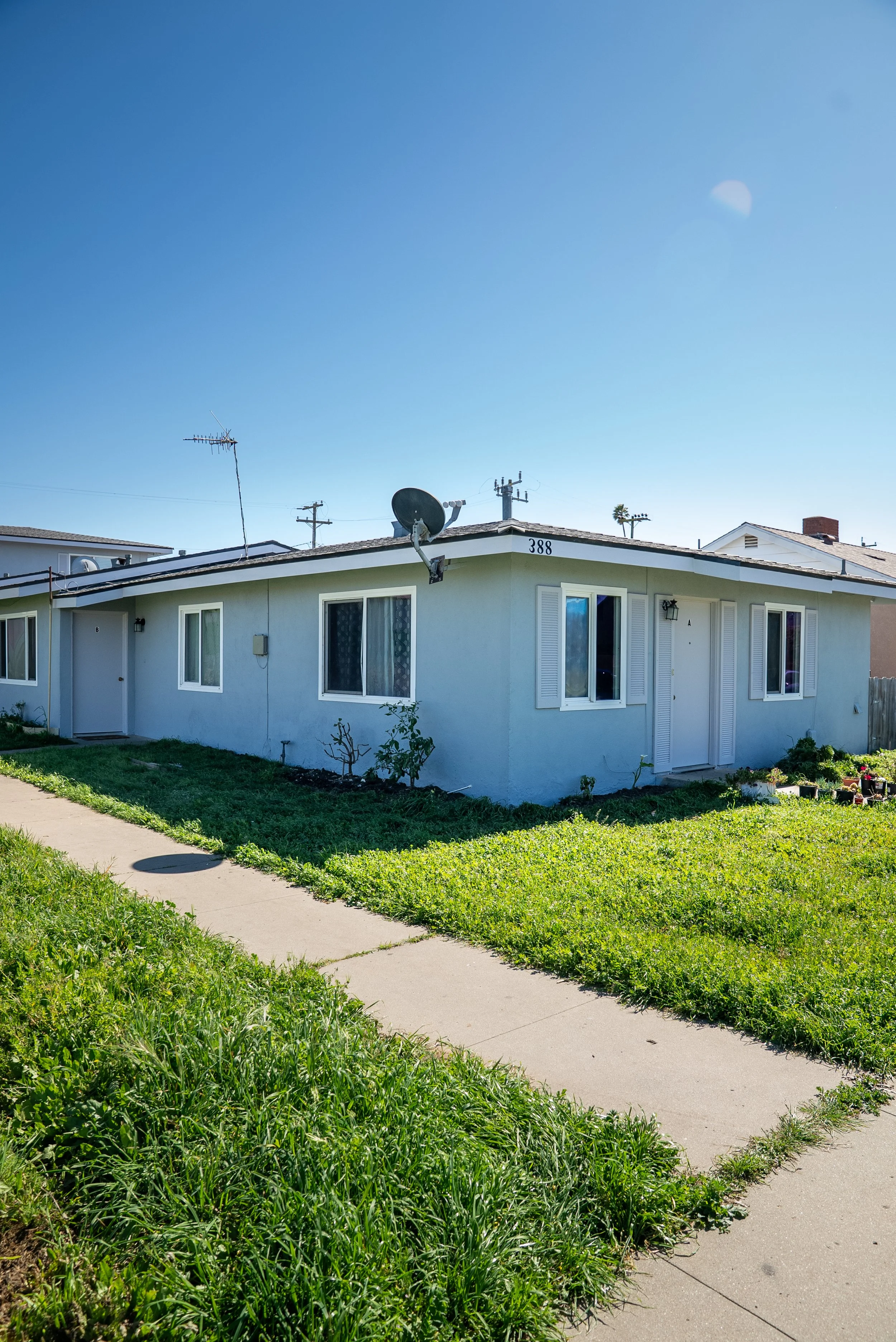 A light blue single-story house with a sloped roof, multiple windows with white shutters, a satellite dish, and an antenna on the roof. A sidewalk runs in front of the house, surrounded by grassy lawn and small plants. The sky is clear and blue.