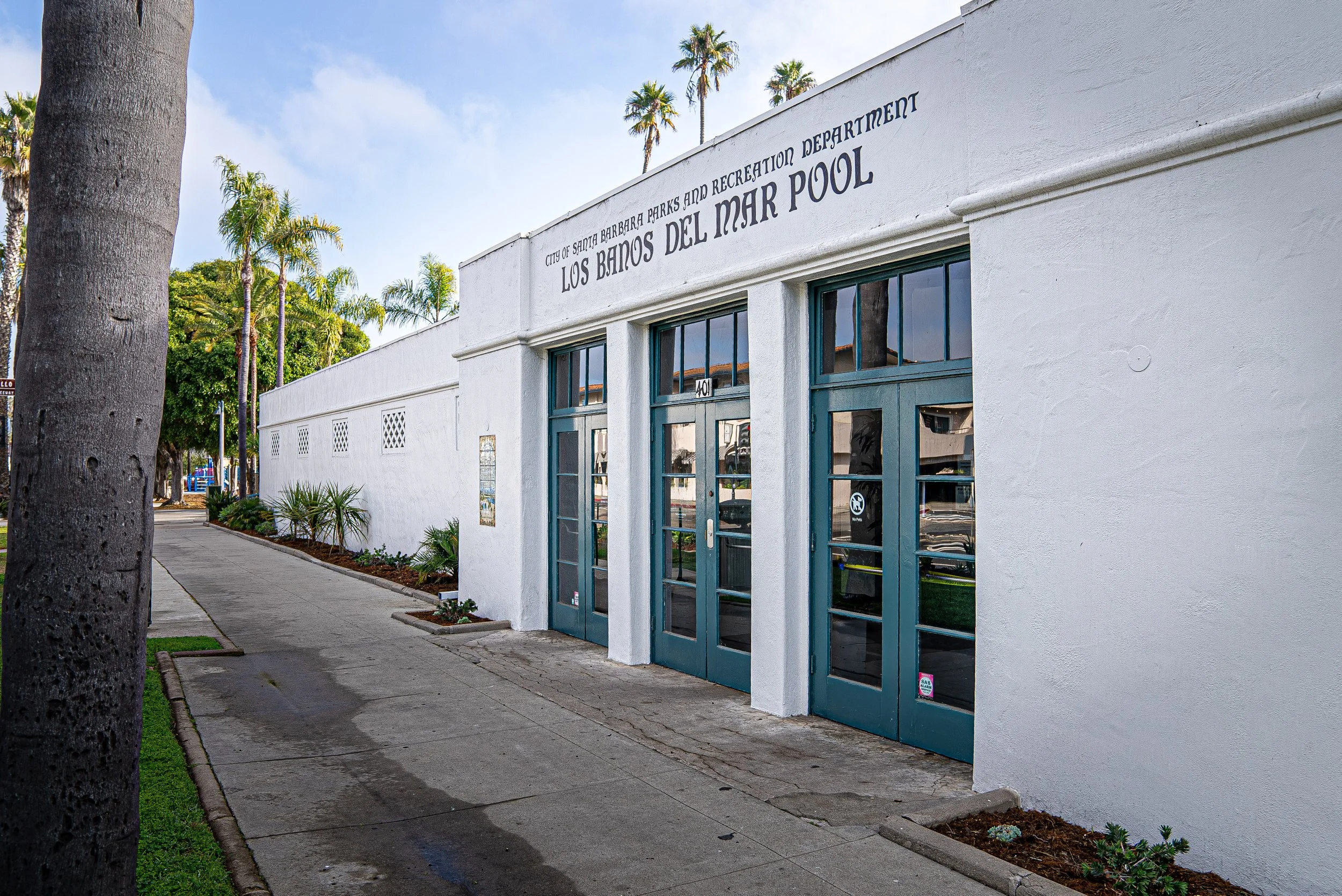 Exterior view of the Los Banos Del Mar Pool building in Santa Barbara, with palm trees and a sidewalk in front.