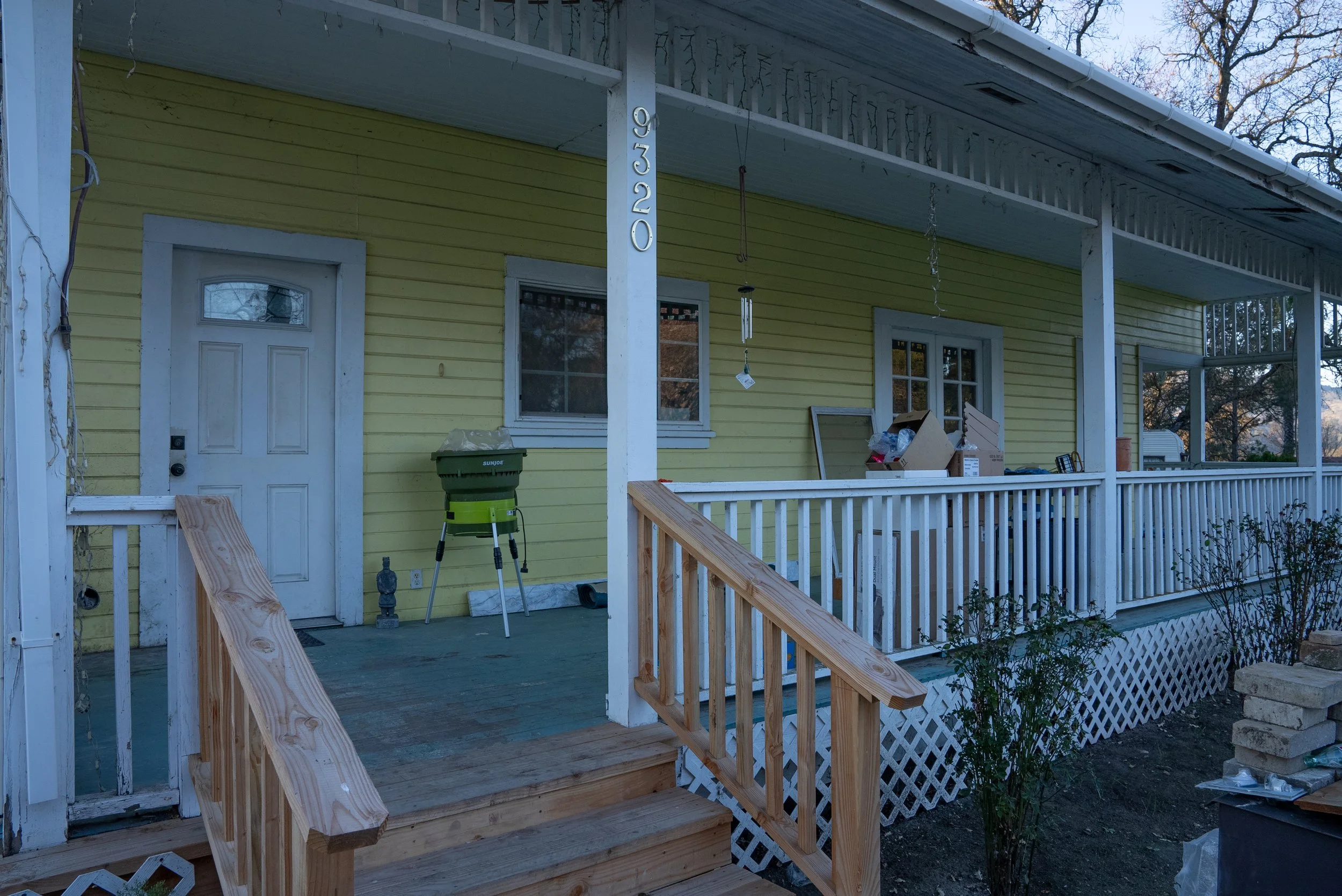 Front porch of a yellow house under construction with new wooden stairs and porch railing, a white door, two windows, a green trash bin, and various construction tools and materials.