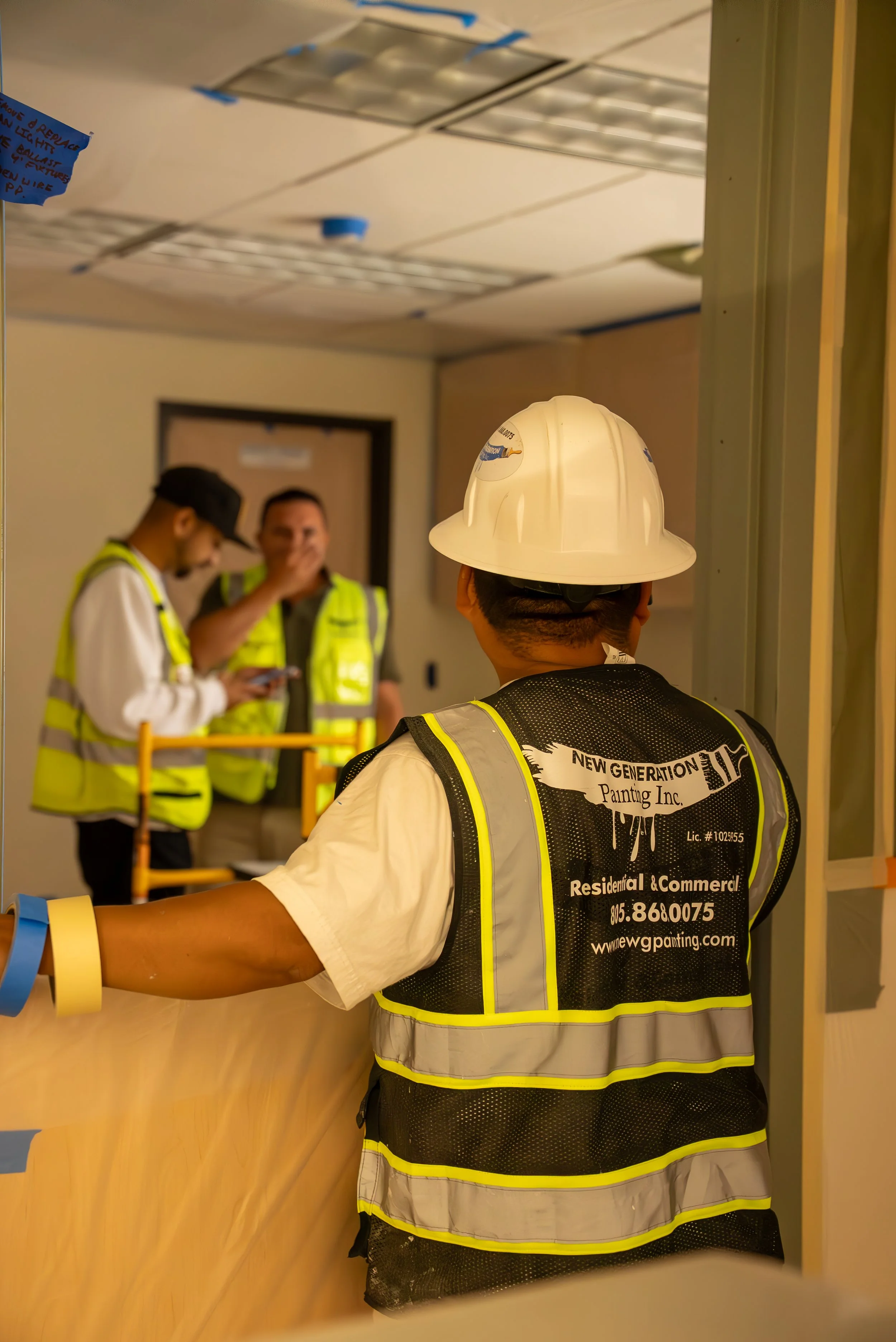 Construction workers wearing yellow safety vests and helmets, standing near a counter in an indoor setting. One worker's vest has company information on the back, including a website and phone number.