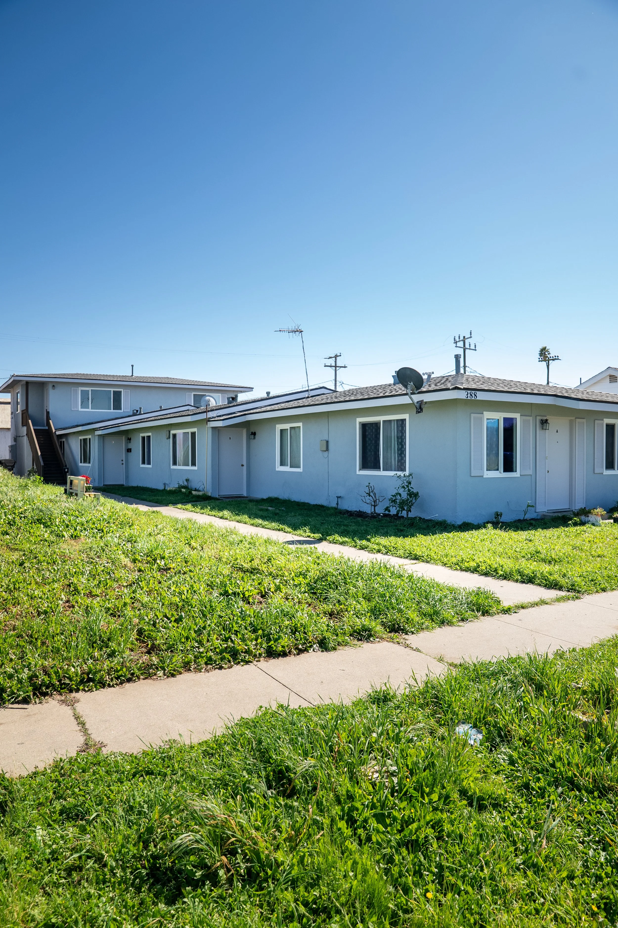 A single-story white residential building with a sloped roof, multiple windows, and a satellite dish on the roof. Green grass and sidewalk in the front yard, with a blue sky overhead.