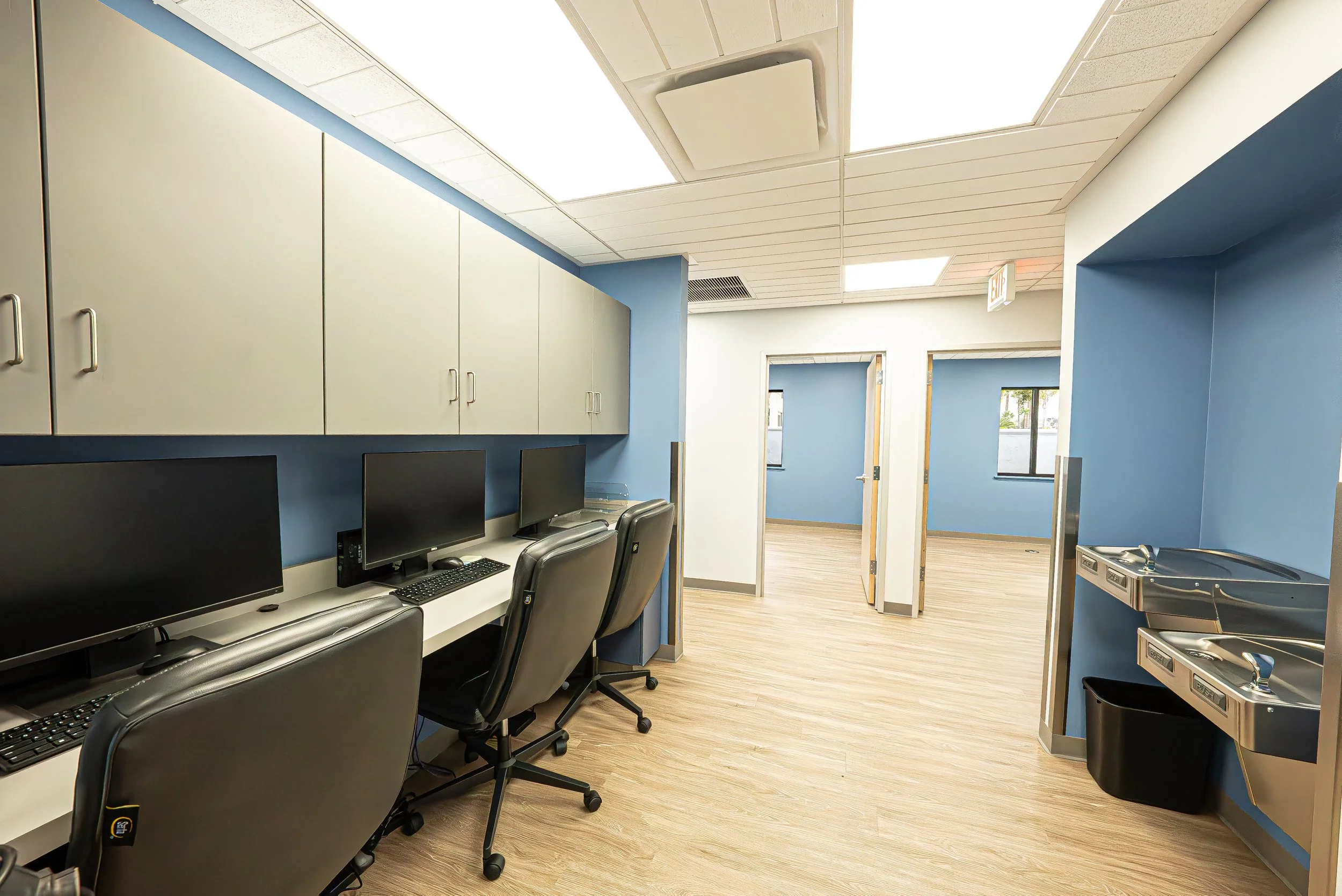 A room with blue walls, white cabinets, two computer workstations with monitors, a sink, and a hallway with doors and windows.