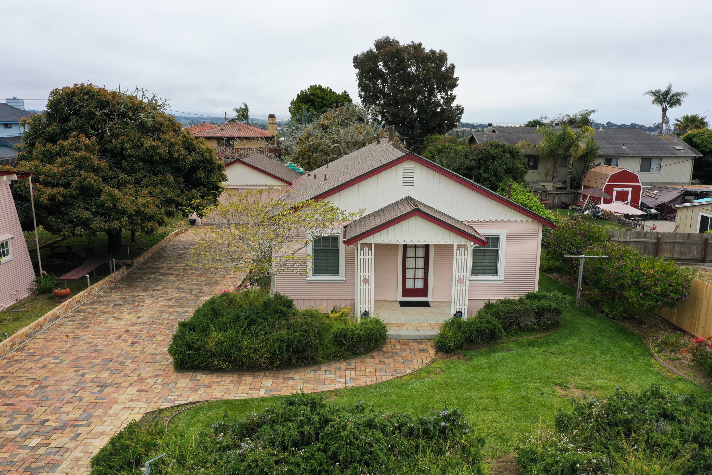 A pink house with white trim, a brick pathway, green bushes, and trees in a residential neighborhood under a cloudy sky.