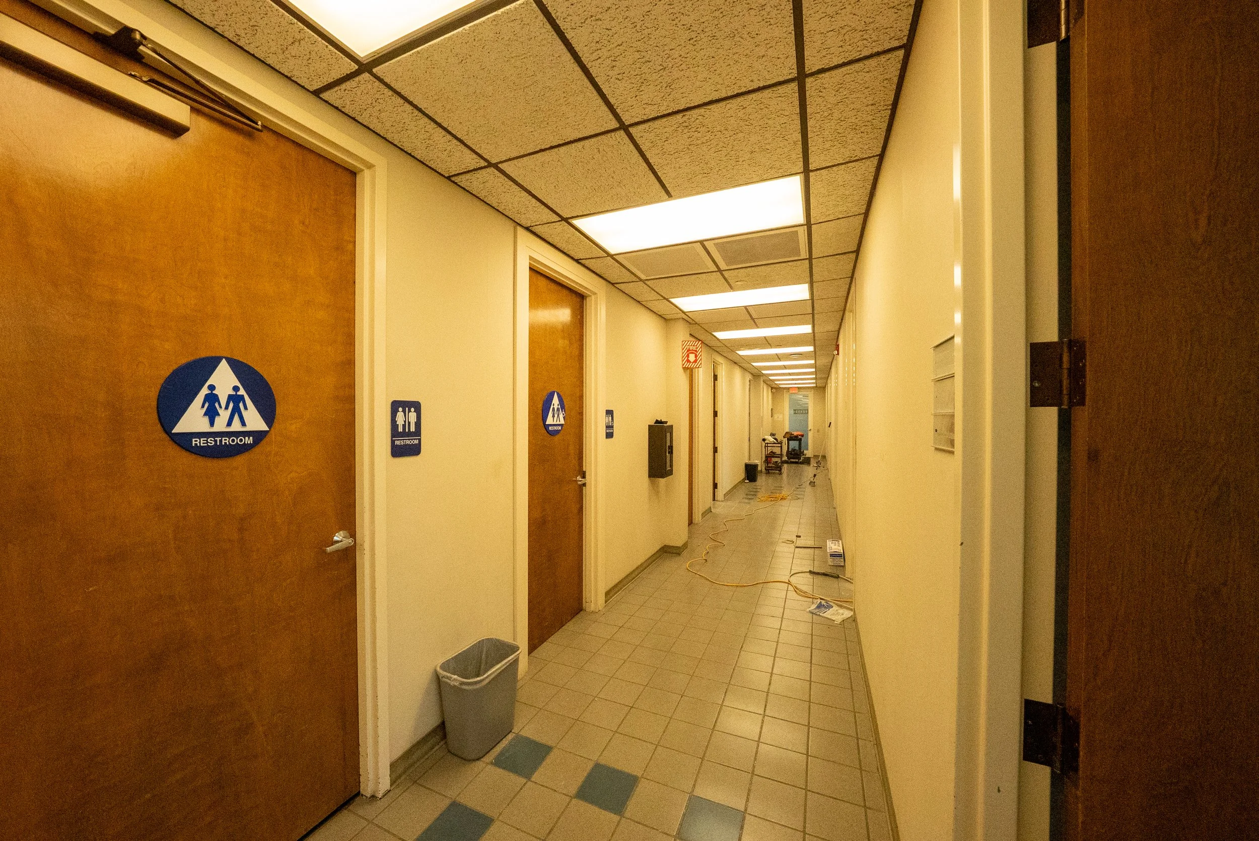Hospital hallway with bathroom signs. Wooden doors on left, beige walls, tiled floor, ceiling with rectangular fluorescent lights. Trash bin seen near the door. Some equipment and cords on floor at the end of the hall.