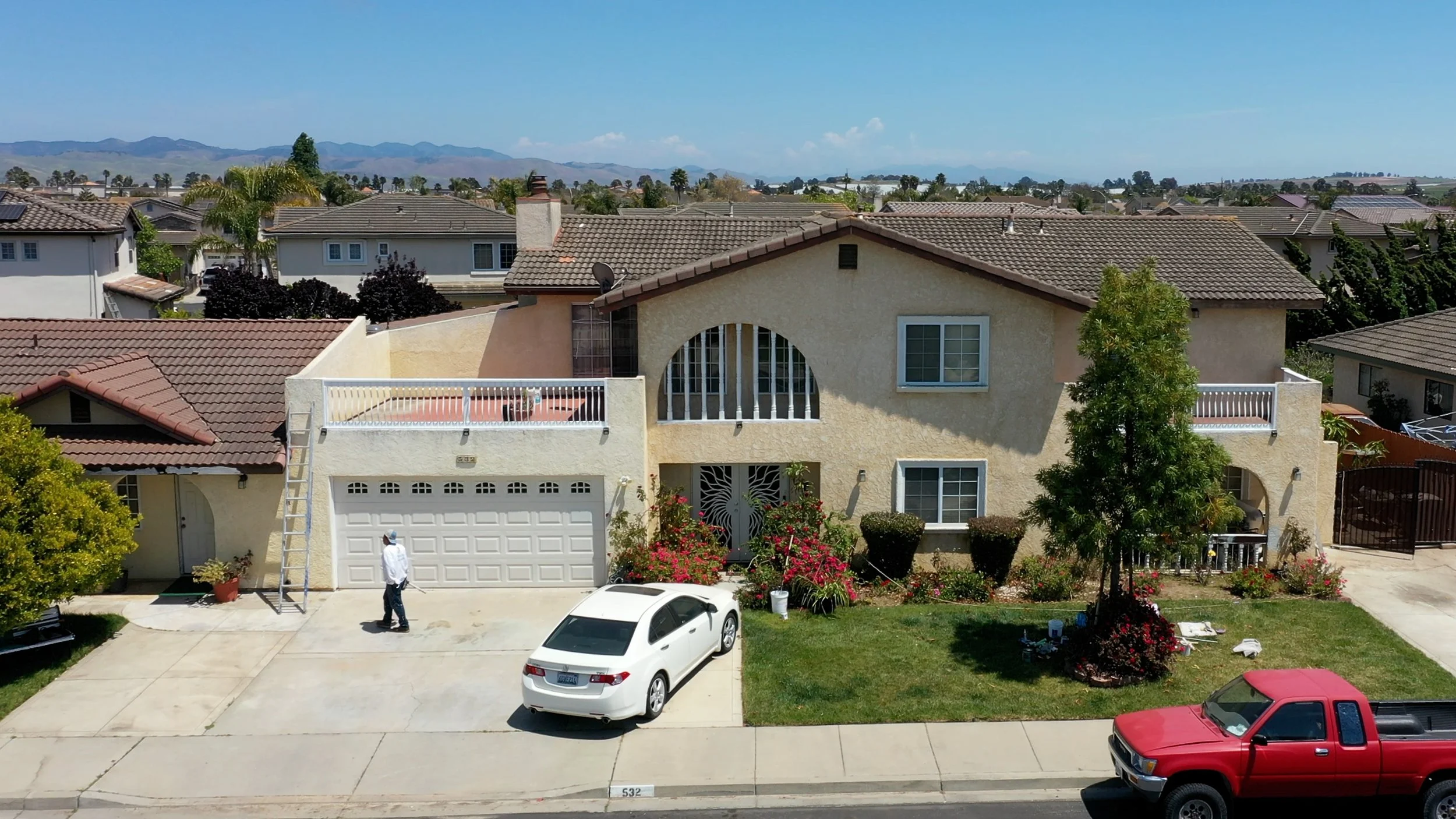 View of a residential neighborhood with a beige two-story house with a garage, a man walking on the driveway, parked cars, and a background of numerous houses and mountains under a clear sky.