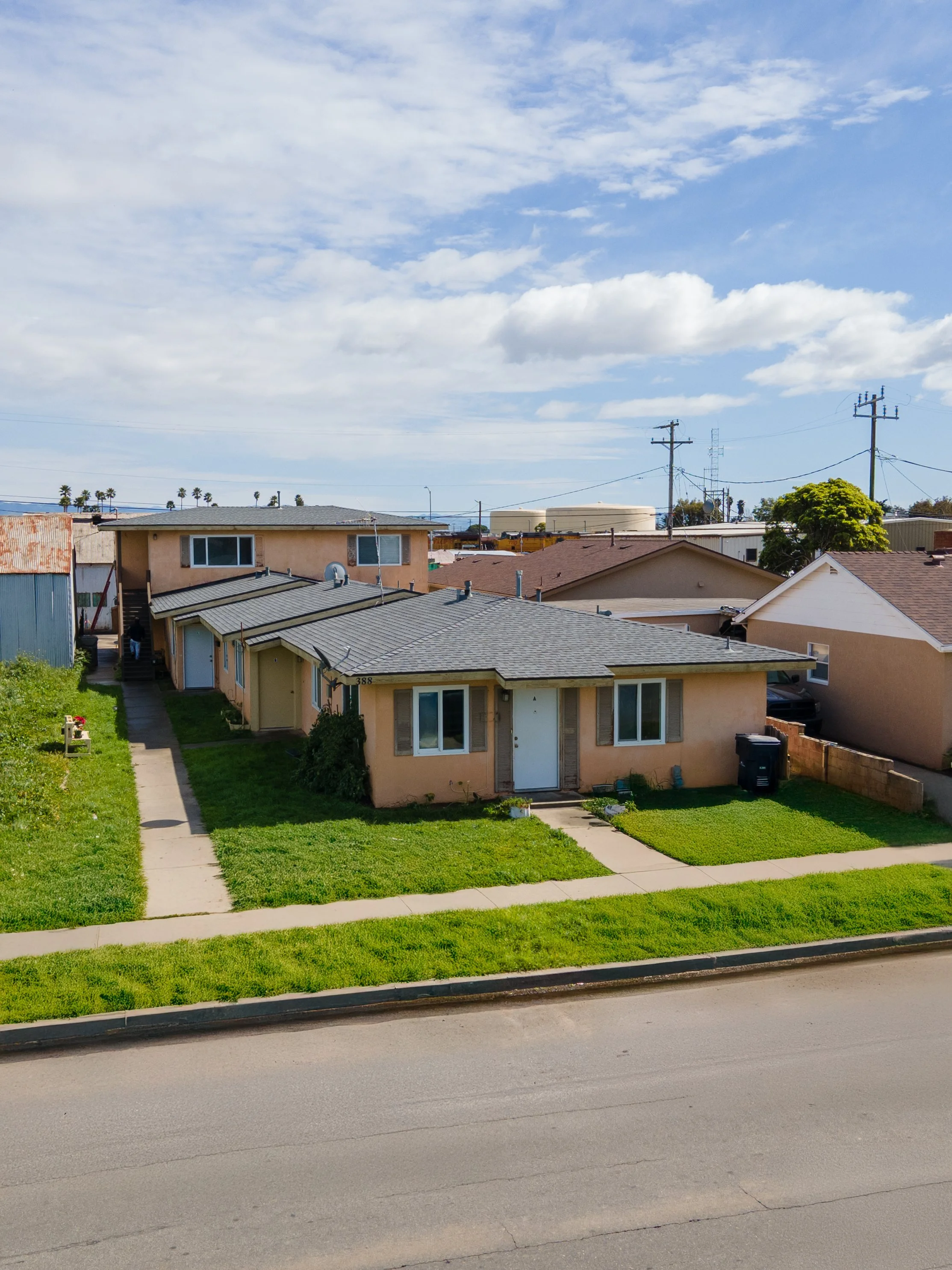 A view of a residential neighborhood with a sidewalk, green grass, and a row of houses with beige and white exteriors. Power lines and utility poles are visible against a partly cloudy sky.