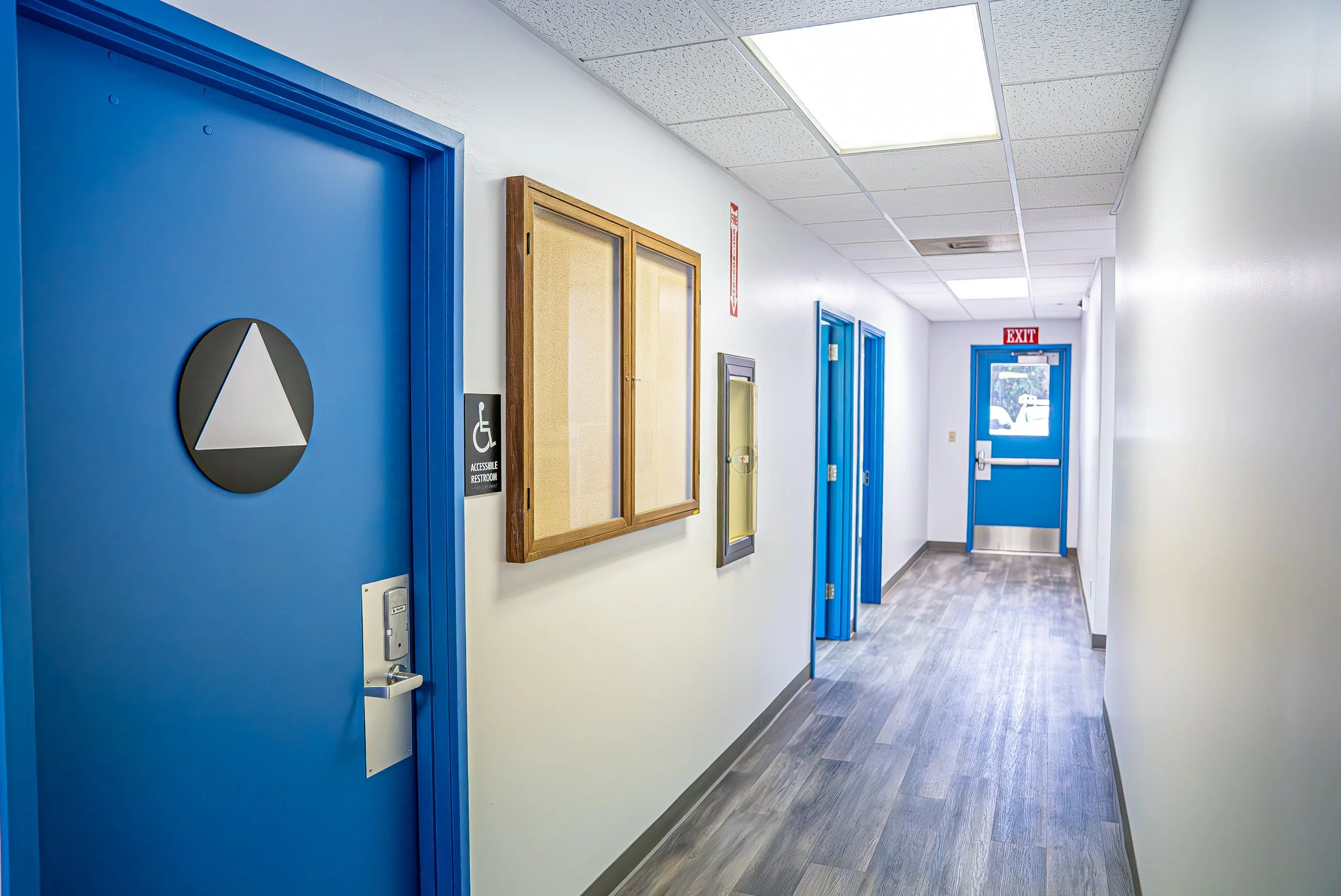 A hallway in a building with white walls, a drop ceiling with fluorescent lights, blue doors, a bulletin board, an exit door, and an accessible restroom sign.