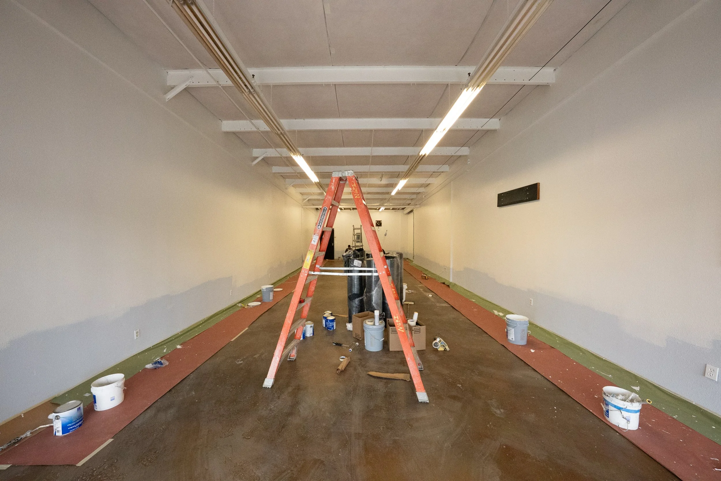A large room under renovation with beige walls, a brown floor, and a drop ceiling. Painting supplies, tools, and an orange step ladder are scattered around. The ceiling has exposed white beams and fluorescent lights. 