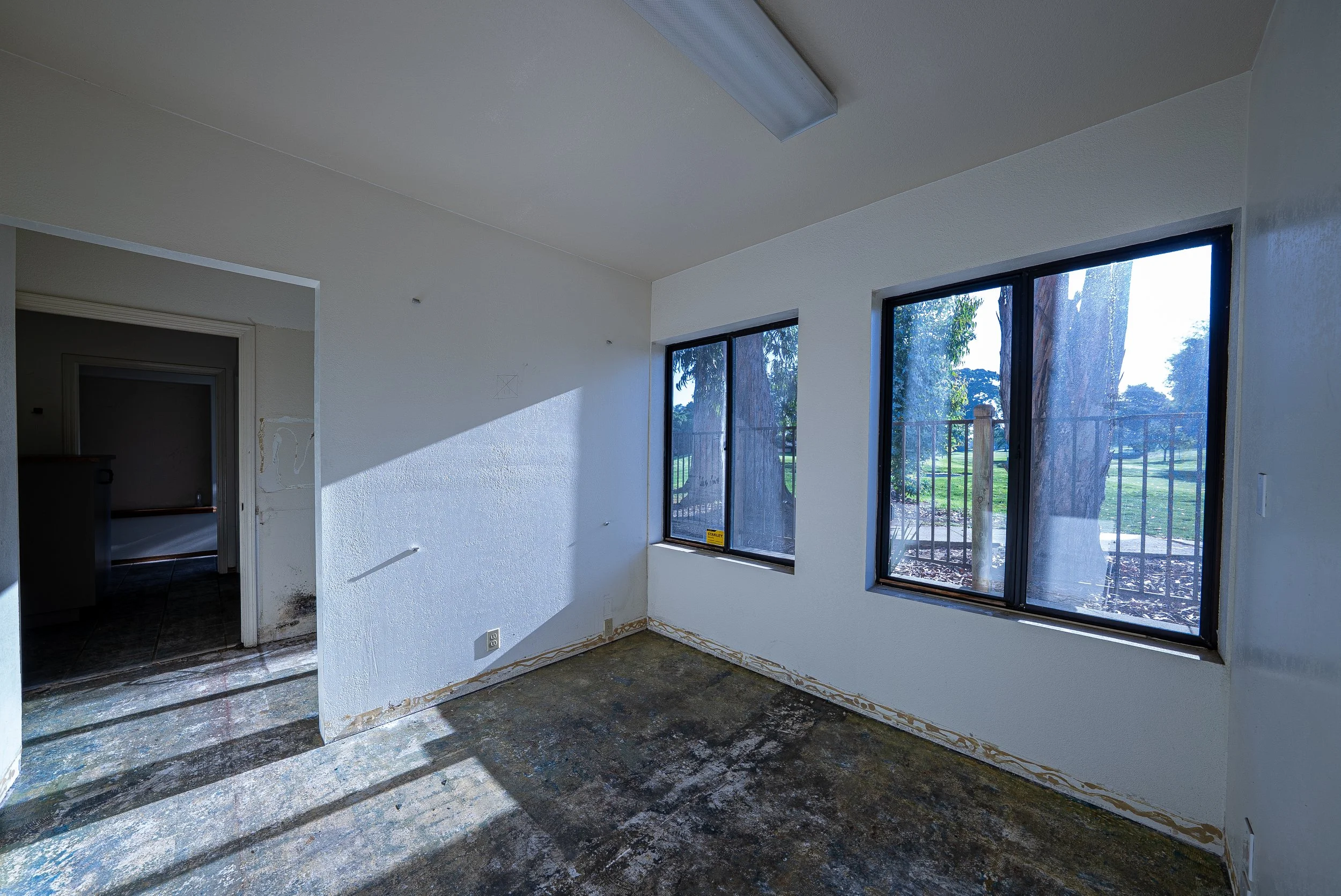 Empty room with large windows showing trees outside, white walls, and a doorway leading to another room. Sunlight casts shadows on the floor.