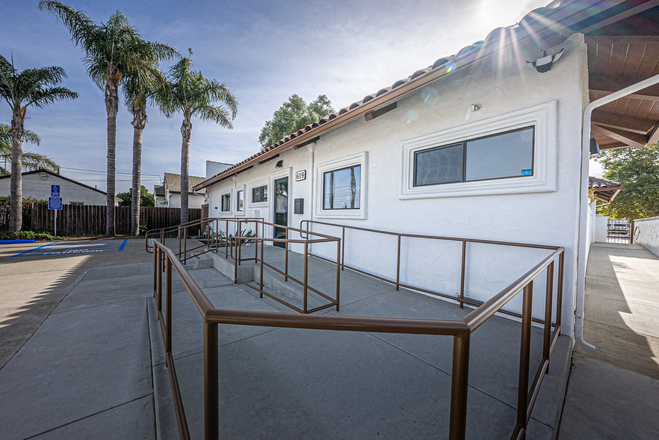 Exterior of a white building with three rectangular windows, palm trees, a parking lot with handicap parking space, and a handicap parking sign, under a clear blue sky with sunlight.