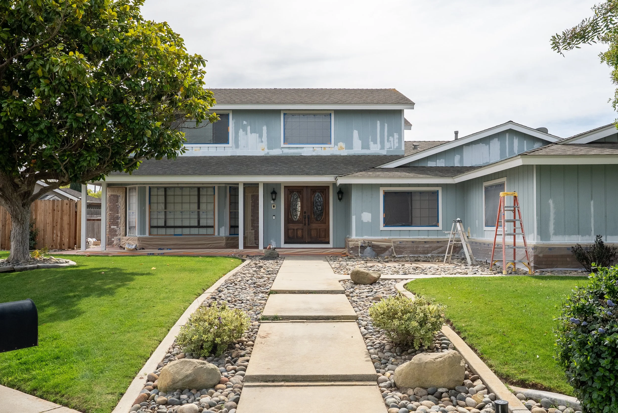 Front yard and house undergoing exterior renovation, with a pathway leading to the front door, a tree on the left, and construction tools on the right.