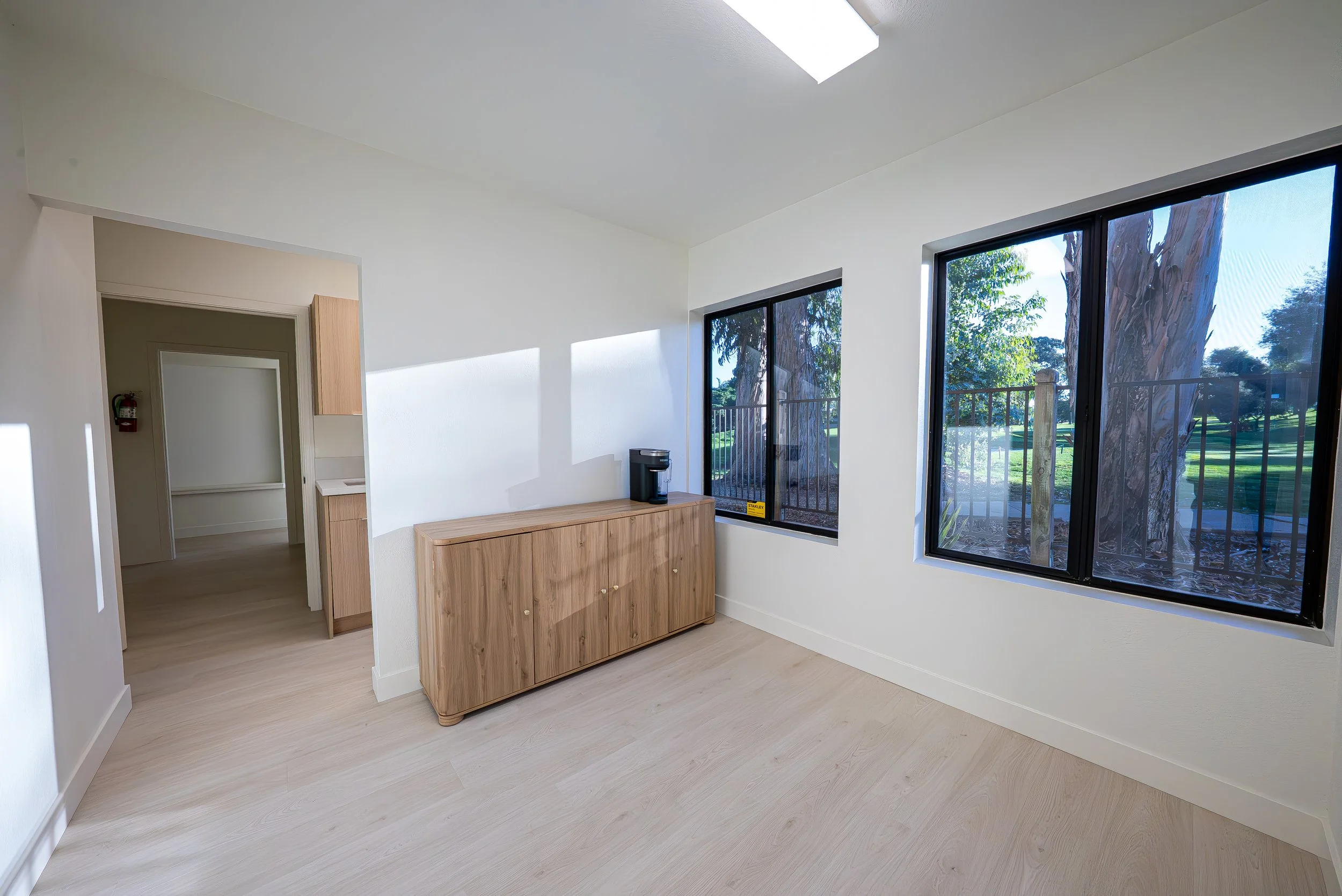 Interior view of a room with white walls, large windows showing trees outside, a wooden cabinet with a black coffee maker, and a doorway leading to another room.