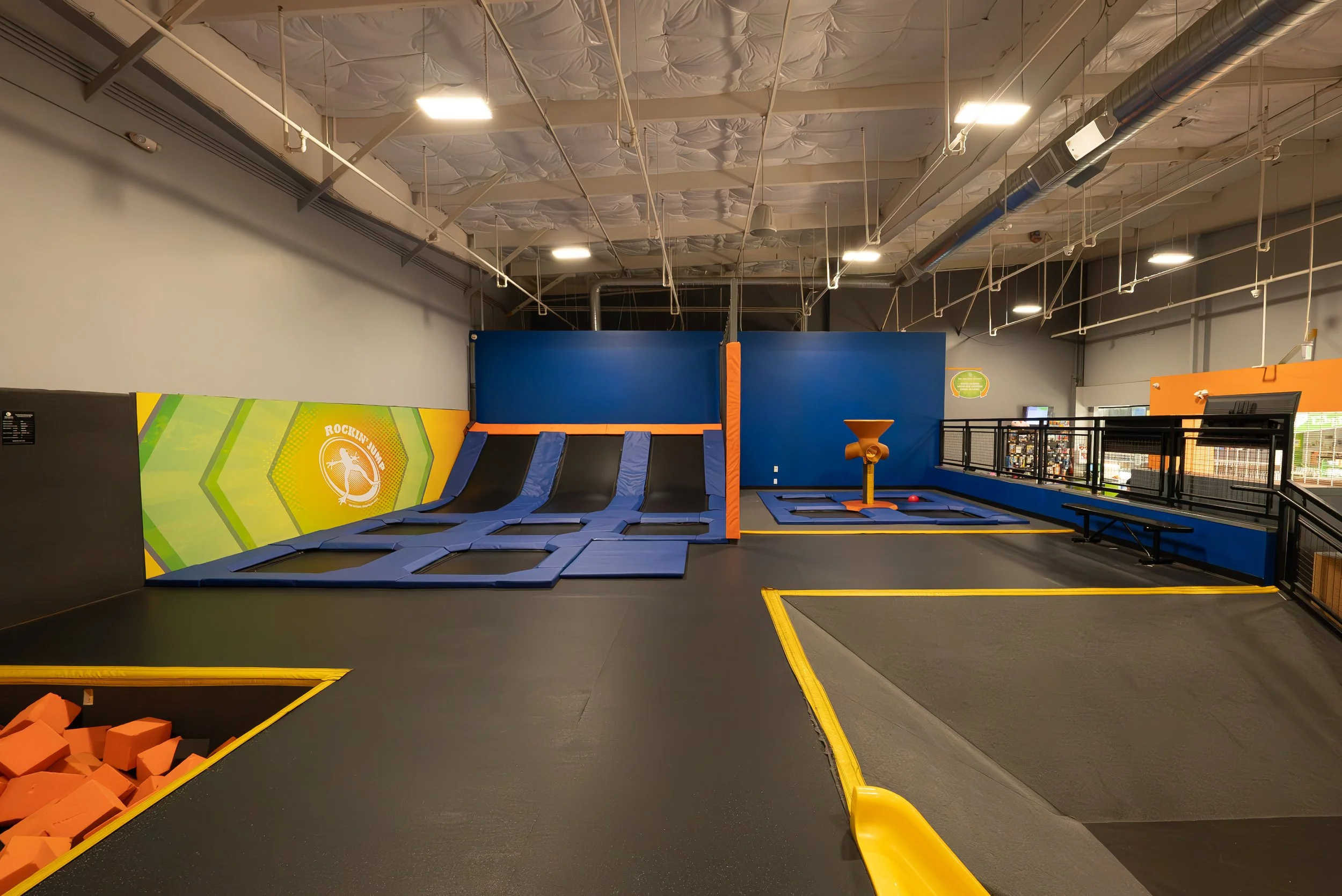 An indoor trampoline park with several intertwined trampolines, foam blocks in orange, and a blue safety wall. The ceiling has exposed ductwork and lighting fixtures.
