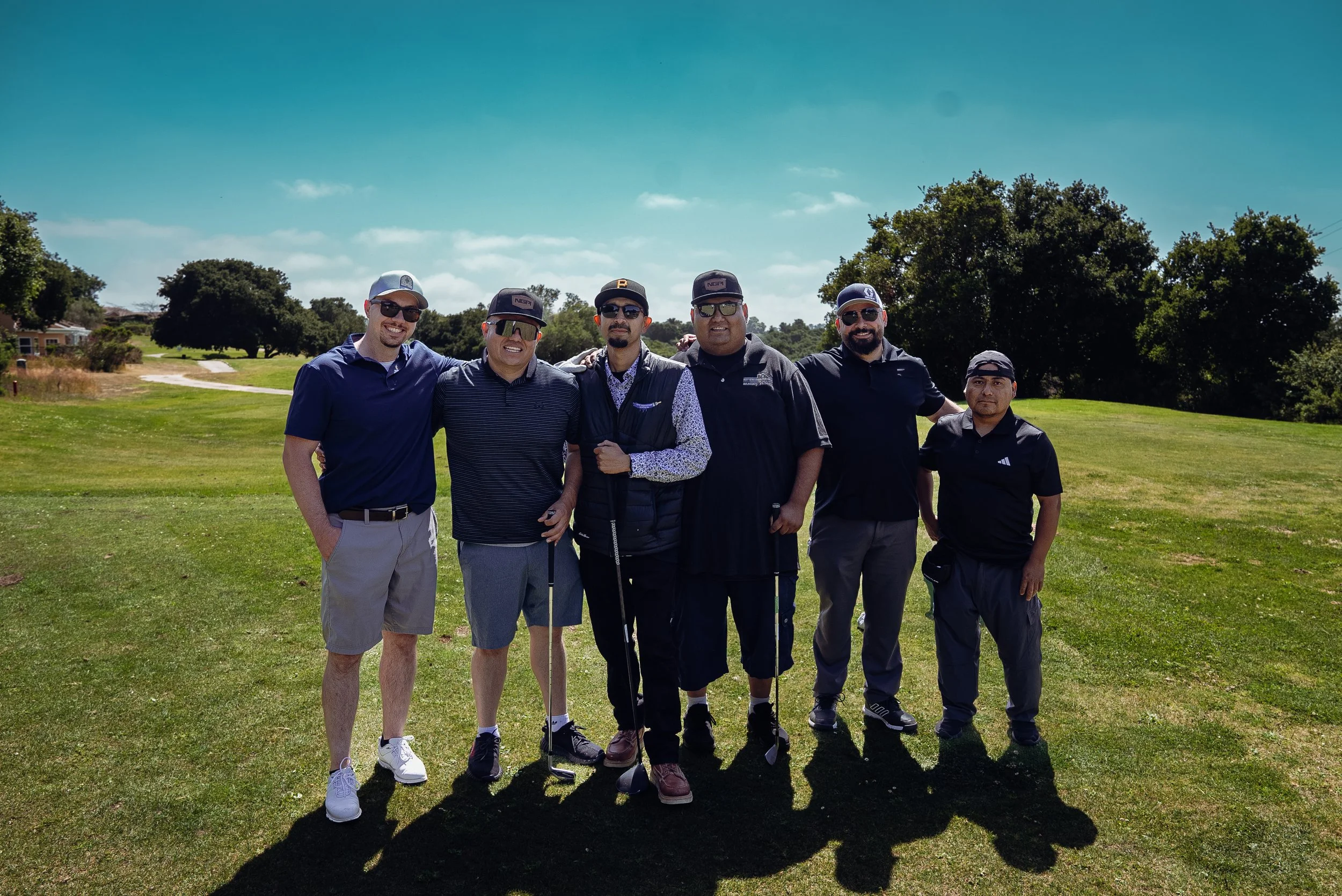 Group of six men standing outdoors on a golf course, dressed in casual and golf attire, smiling at the camera on a sunny day with trees and houses in the background.