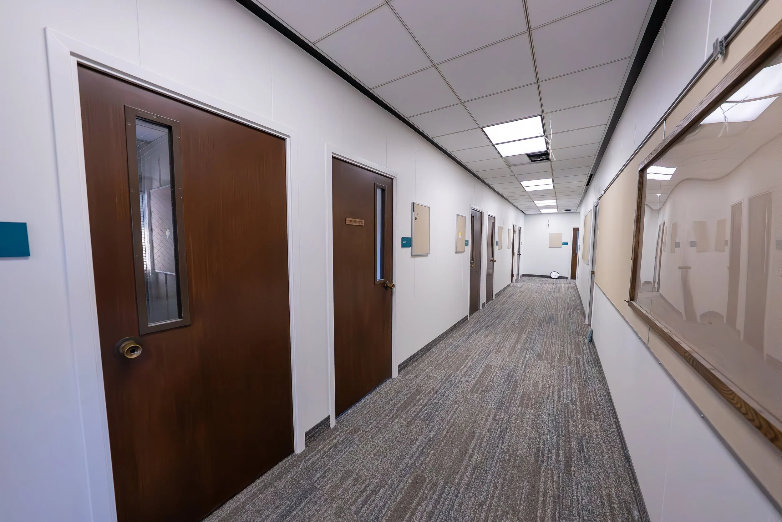 A hallway of an office or medical building with multiple closed wooden doors and framed artwork on white walls. The ceiling has fluorescent lights and a missing panel. The floor is covered with a patterned carpet.