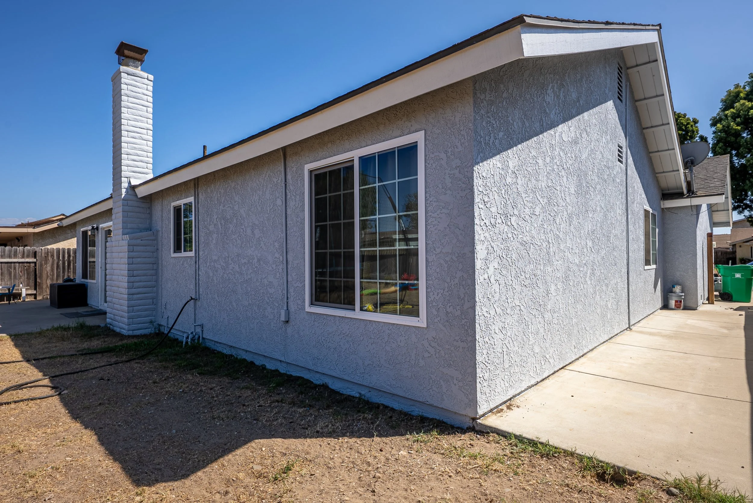 Back view of a white stucco house with a large window, a chimney, a satellite dish, and a side door. The yard has dirt and patches of grass with a concrete patio area.