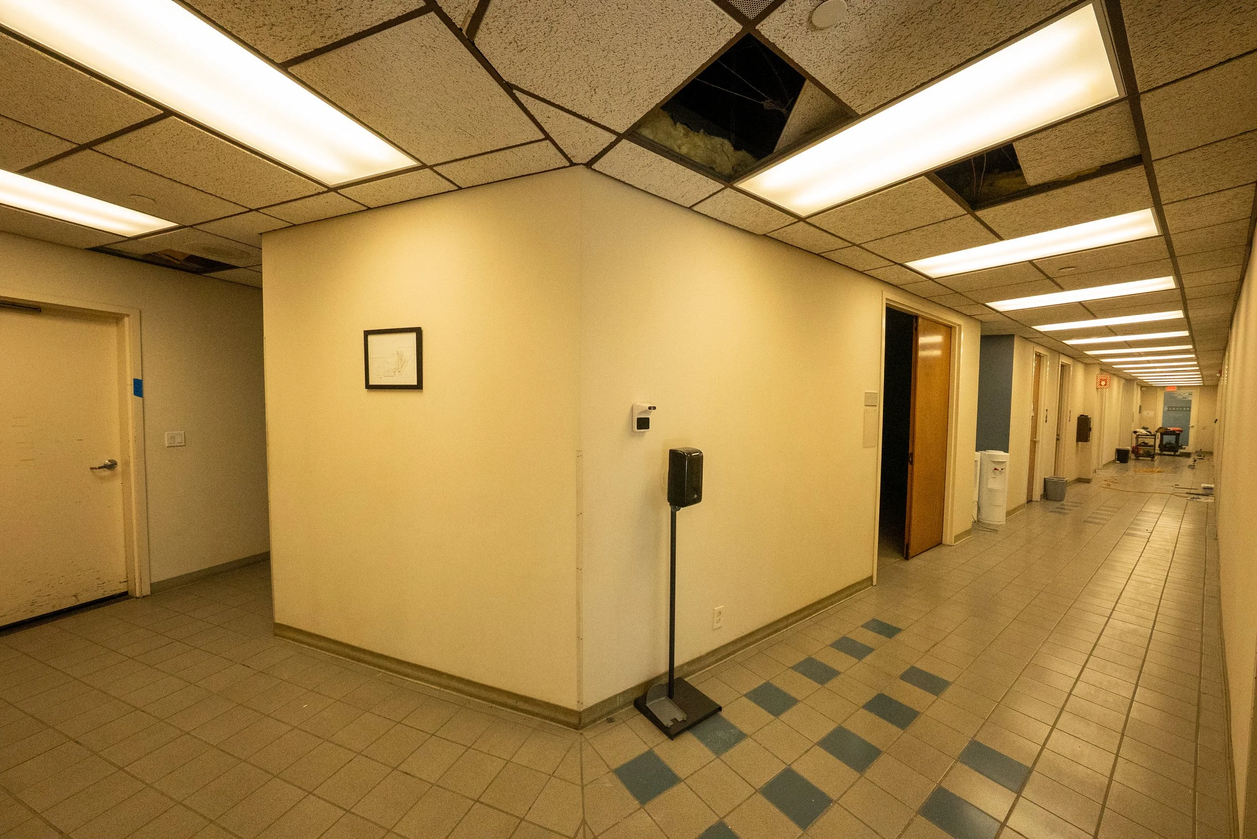 Office hallway with beige walls, ceiling tiles, and fluorescent lighting. One ceiling tile is missing, exposing the ceiling mechanics. The floor has beige and blue tiles, and there is a framed picture on the wall, a hand sanitizer dispenser, a black 