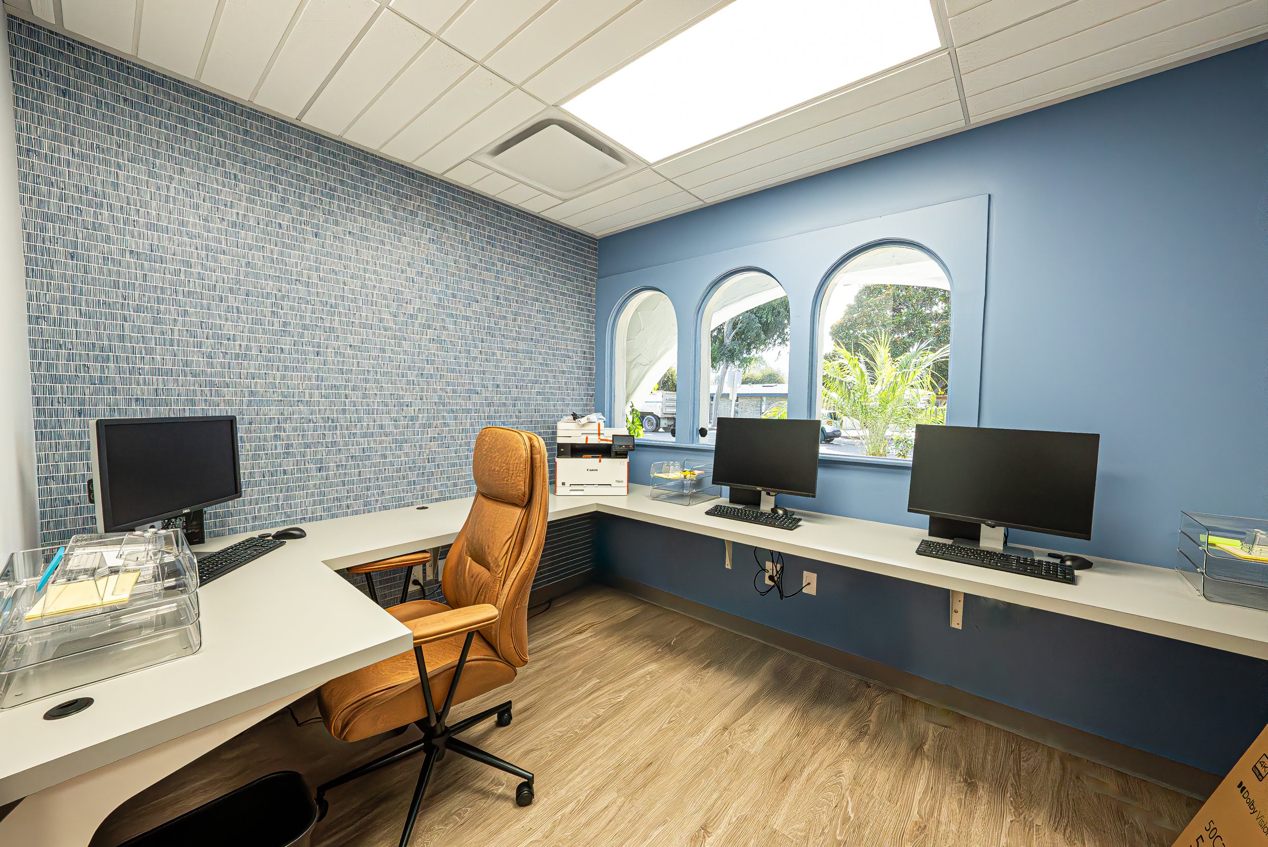 An office with a white desk, four computer monitors, a brown office chair, and a blue accent wall with three windows showing outside greenery.