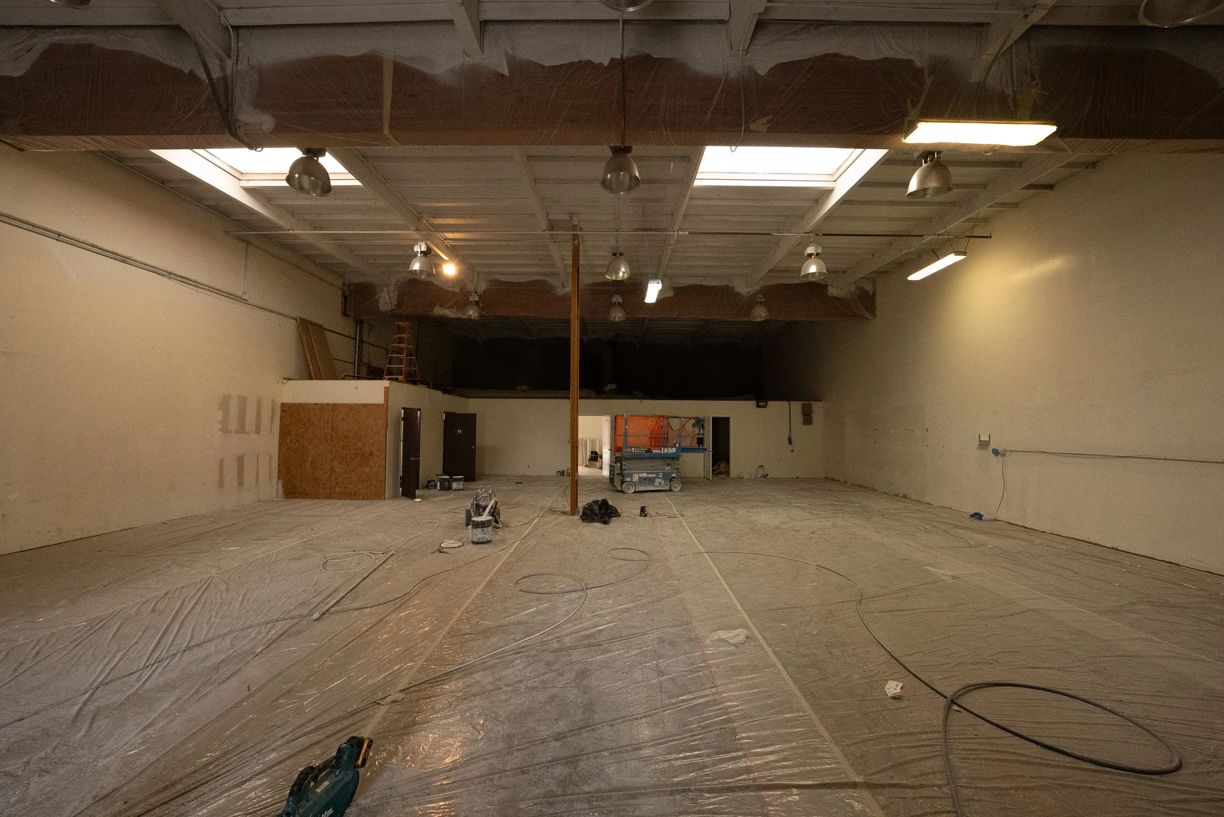 Large indoor space under construction or renovation, with plastic sheeting covering the floor, multiple light fixtures hanging from the ceiling, and tools and equipment scattered around.