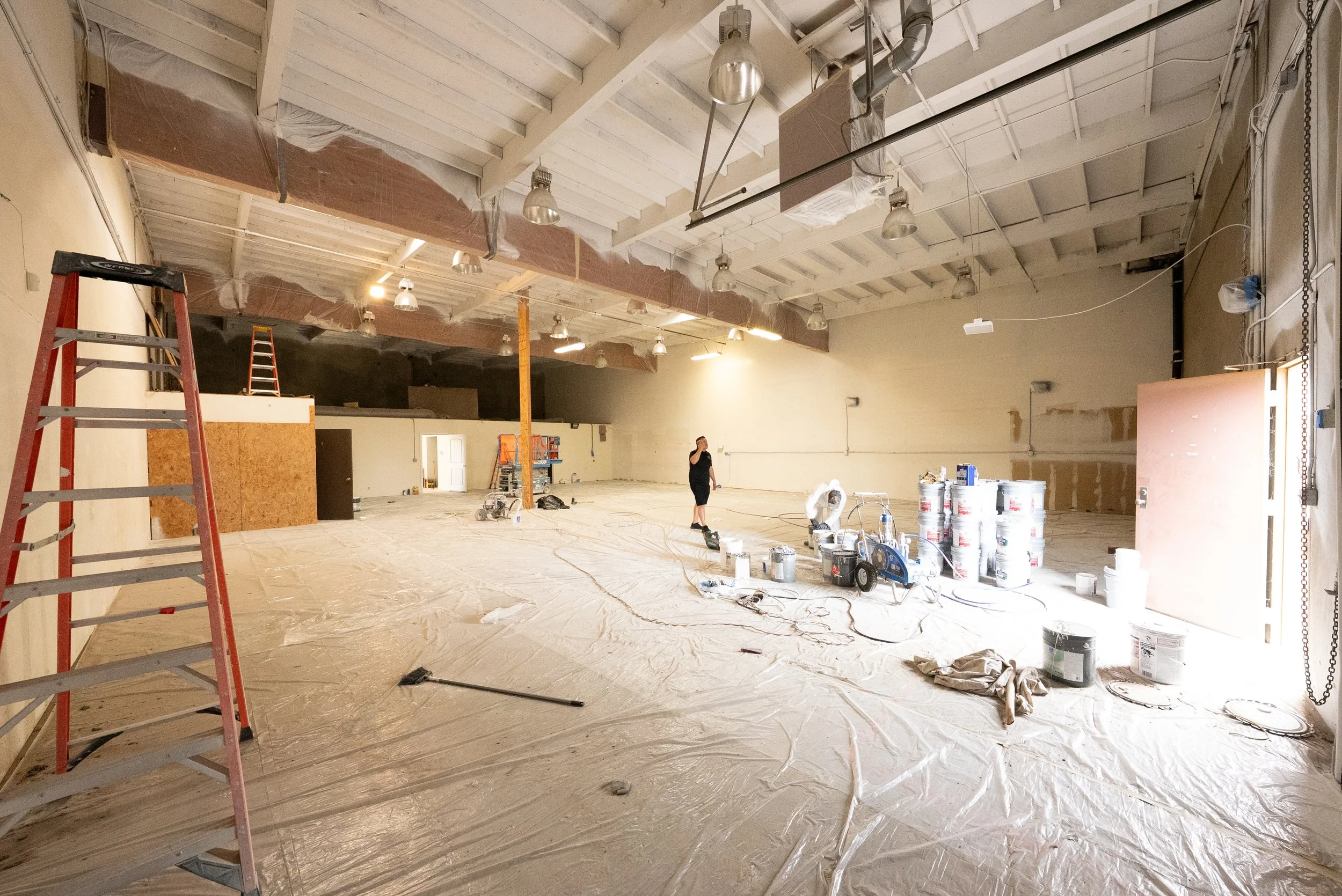 An indoor construction site with a high ceiling, unfinished walls, and scattered construction tools and materials. Ladders, paint cans, and protective covering on the floor are visible, with a person working in the background.