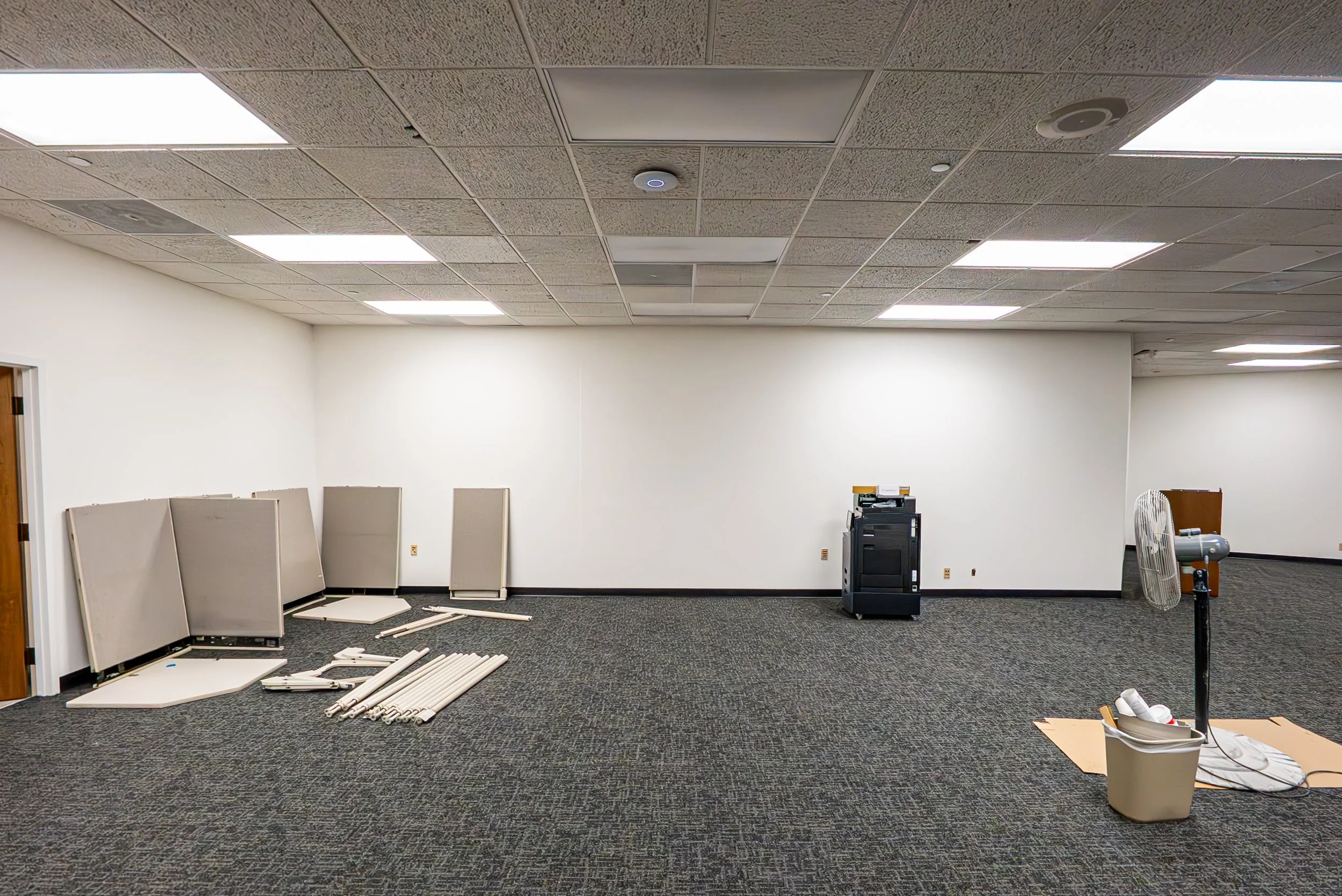 An indoor room with a collapsed drop ceiling, scattered disassembled white panels, pipes, a fan, a black cart, and a trash bin with cords on the floor.