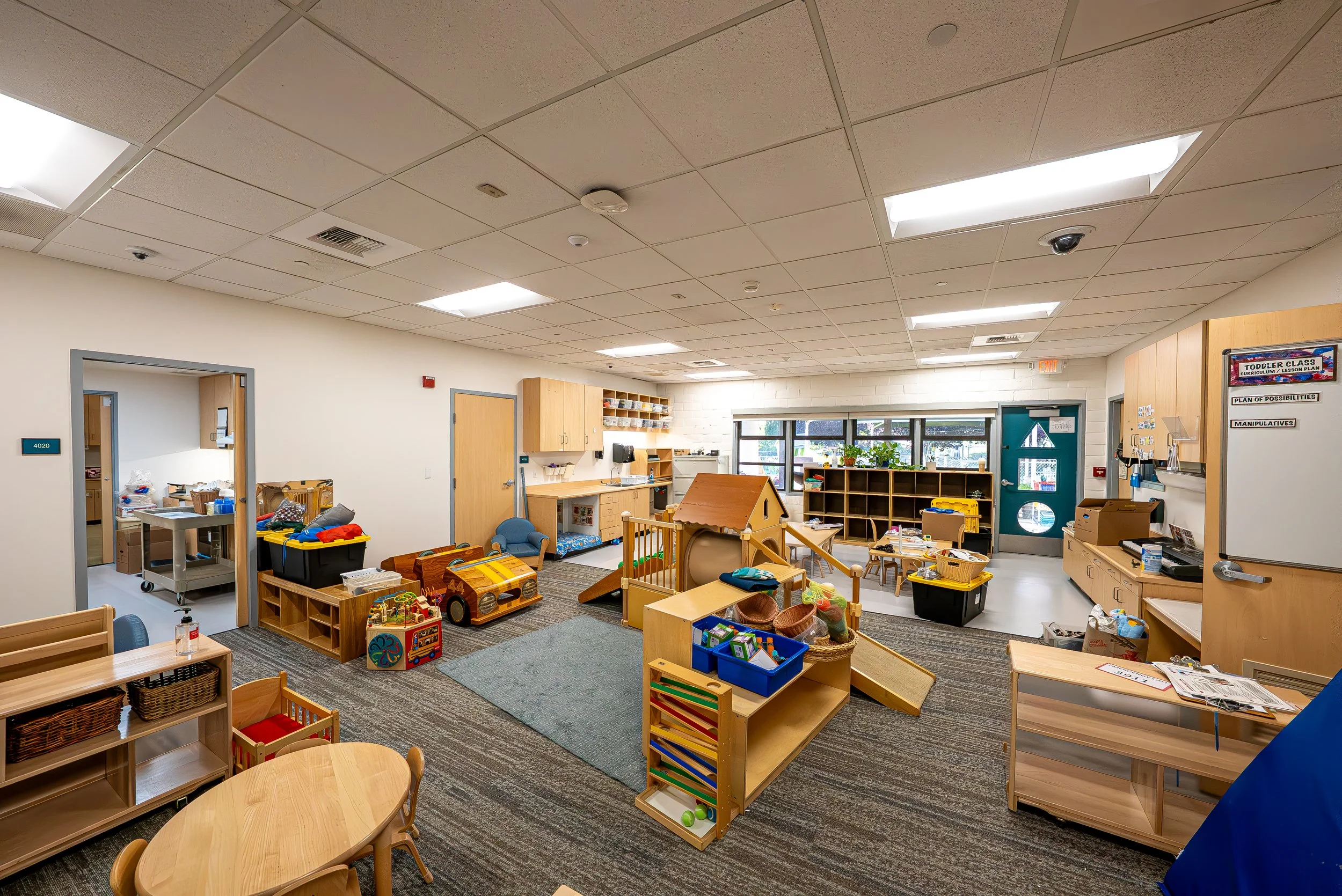 An indoor classroom or playroom filled with toys, small tables, and chairs suitable for young children, with shelves and storage bins, windows letting in natural light, and a colorful, organized environment.