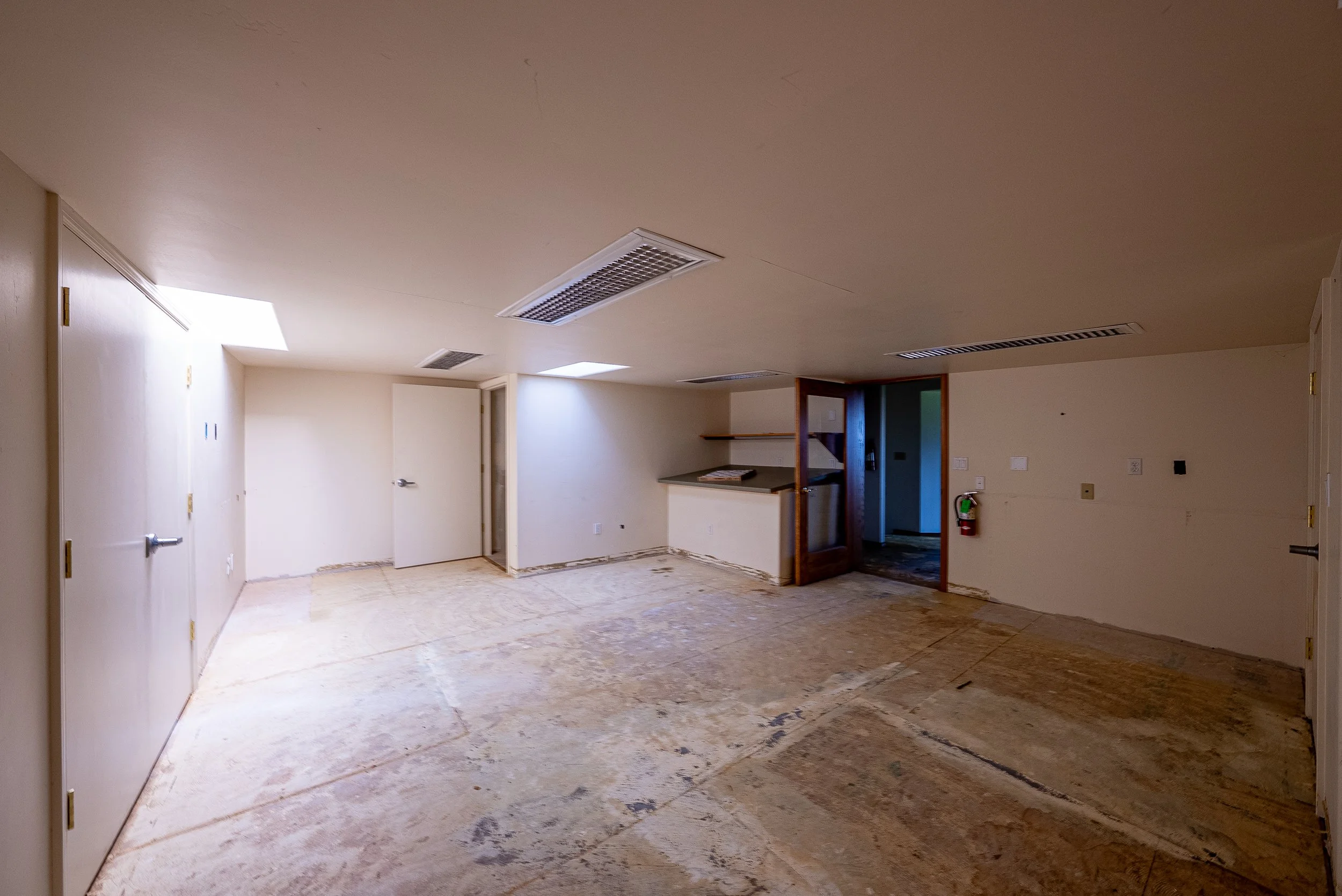 An empty room with a partially finished floor, white walls, and a ceiling with multiple air vents. There are doors and a small opening to another area with a countertop visible.