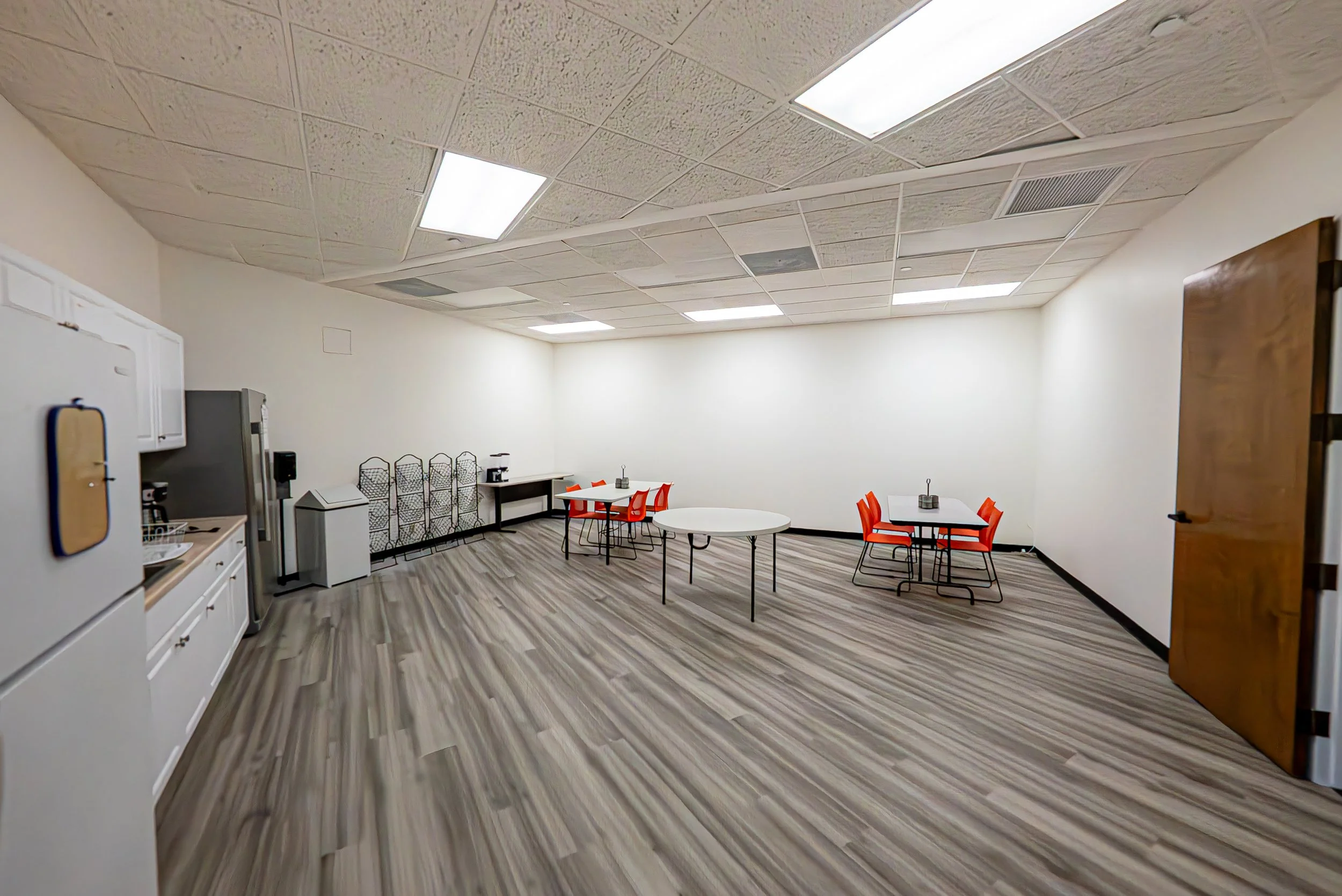 Empty room with tables and chairs, white walls, and a textured ceiling with fluorescent lights.