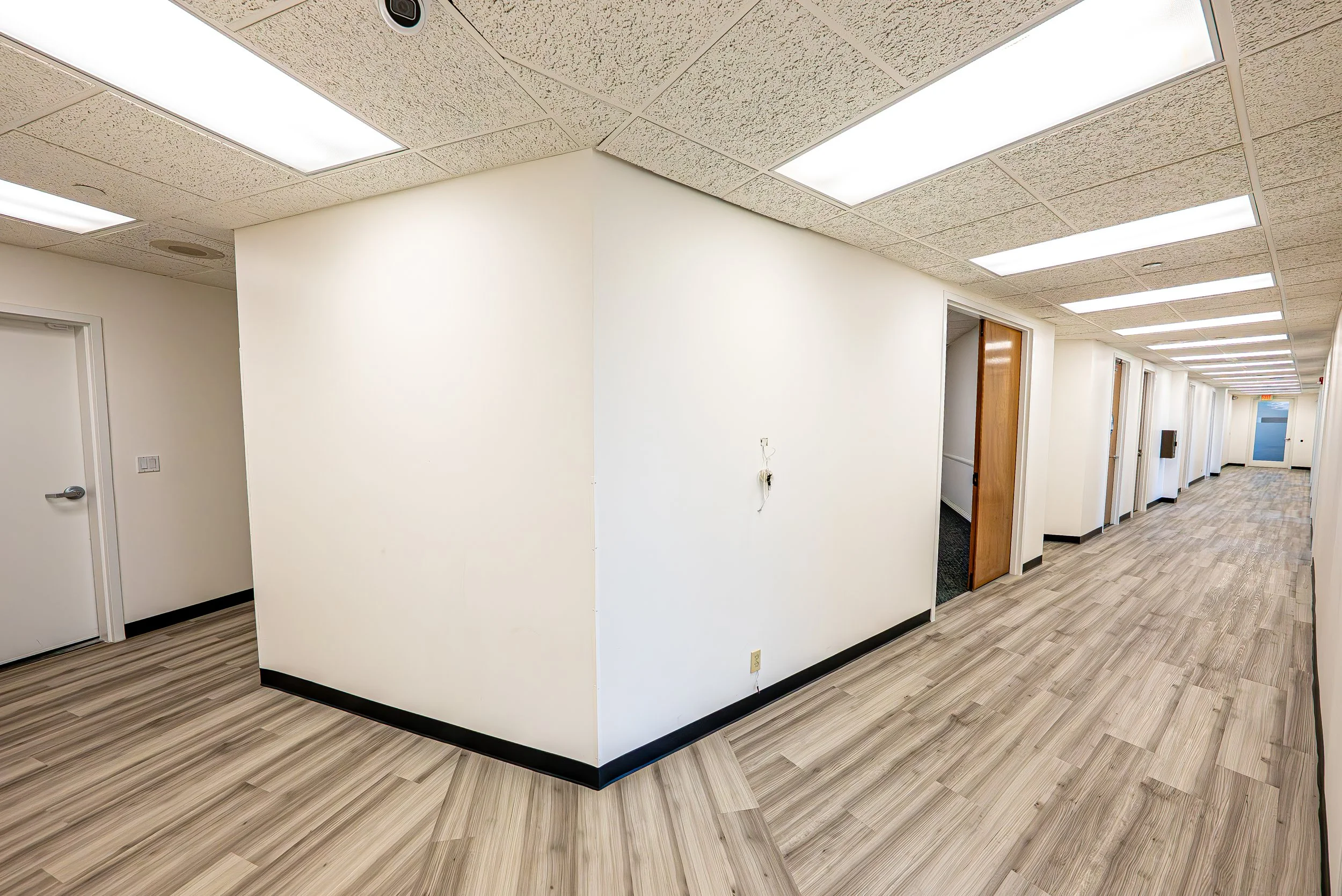 Empty hallway with white walls, wood laminate flooring, ceiling-mounted fluorescent lights, and closed doors.