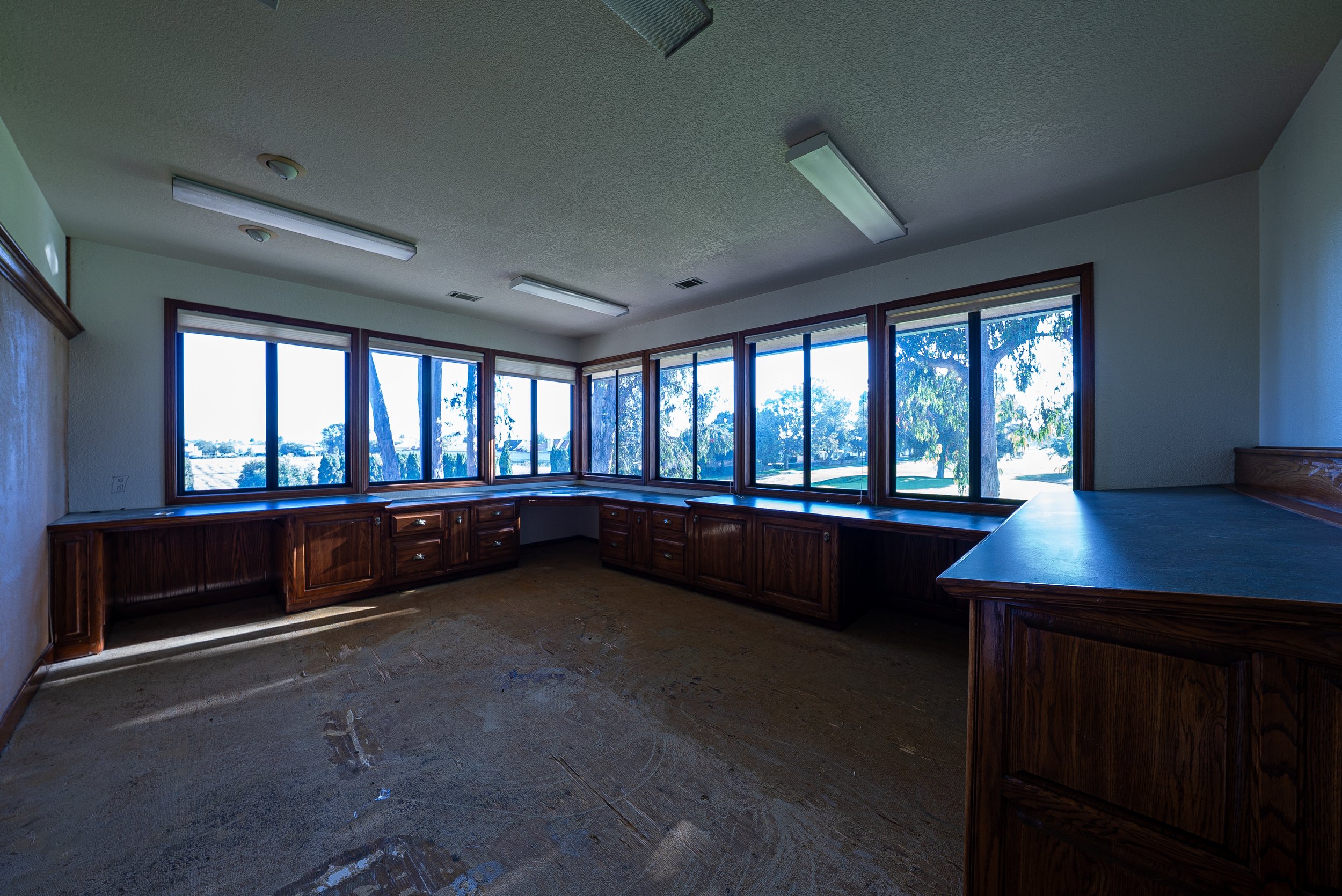 Empty room with large windows, wooden cabinets, and fluorescent ceiling lights.