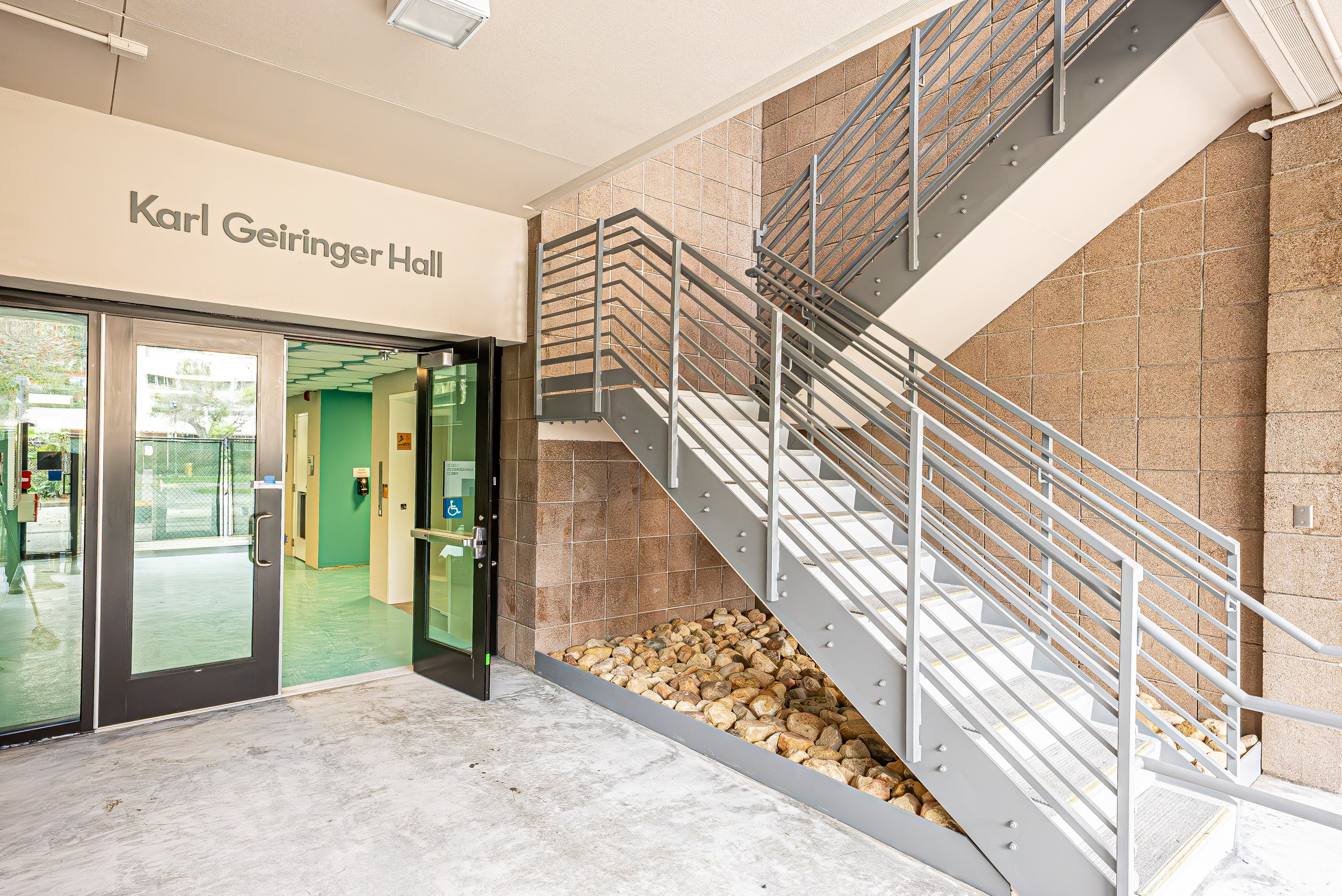 Outside entrance to Karl Geiring Hall with glass doors, metal elevator and external staircase, beige brick wall, and gravel decorative rocks.