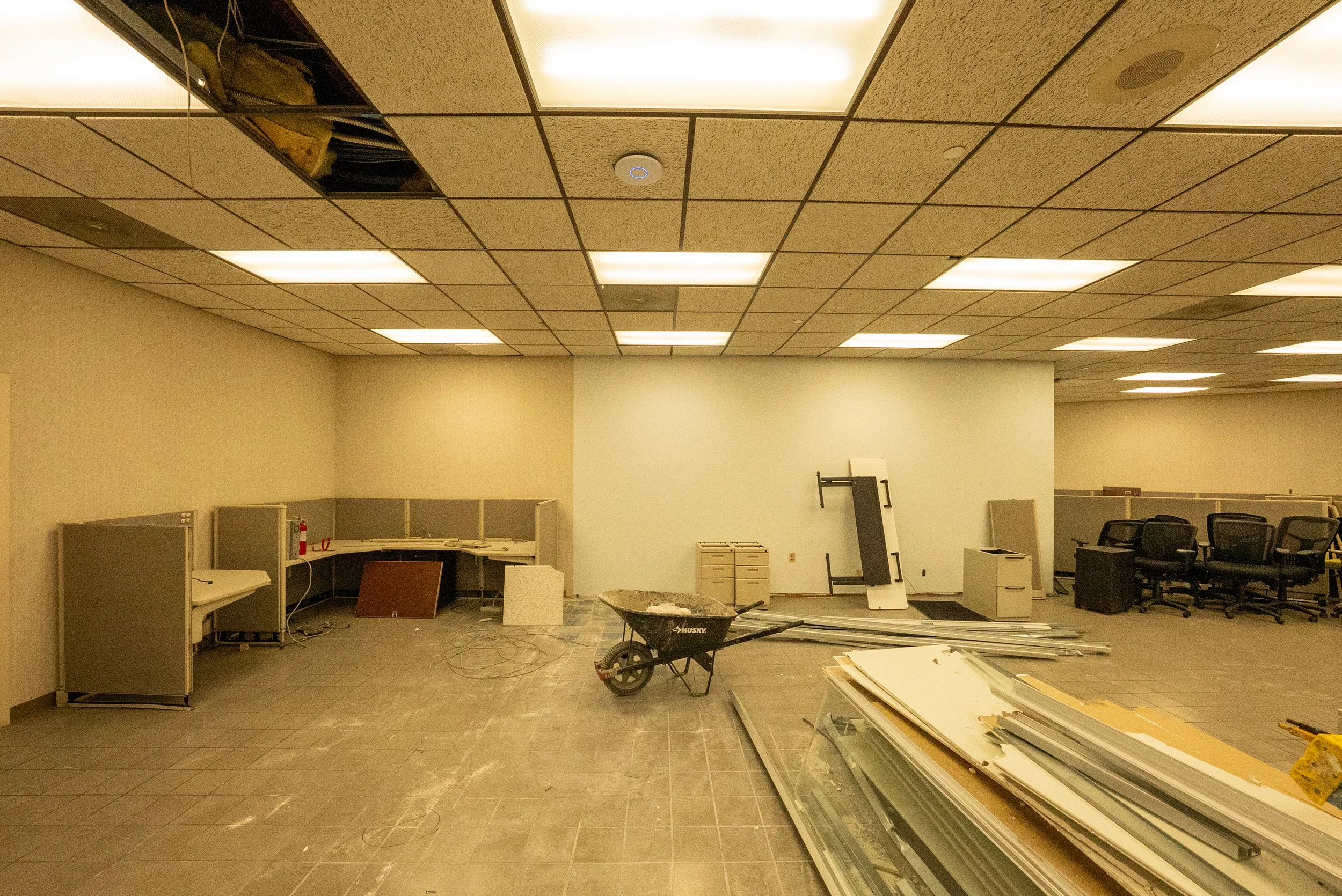 Office space under renovation with ceiling tiles removed, construction materials, and chairs scattered around.